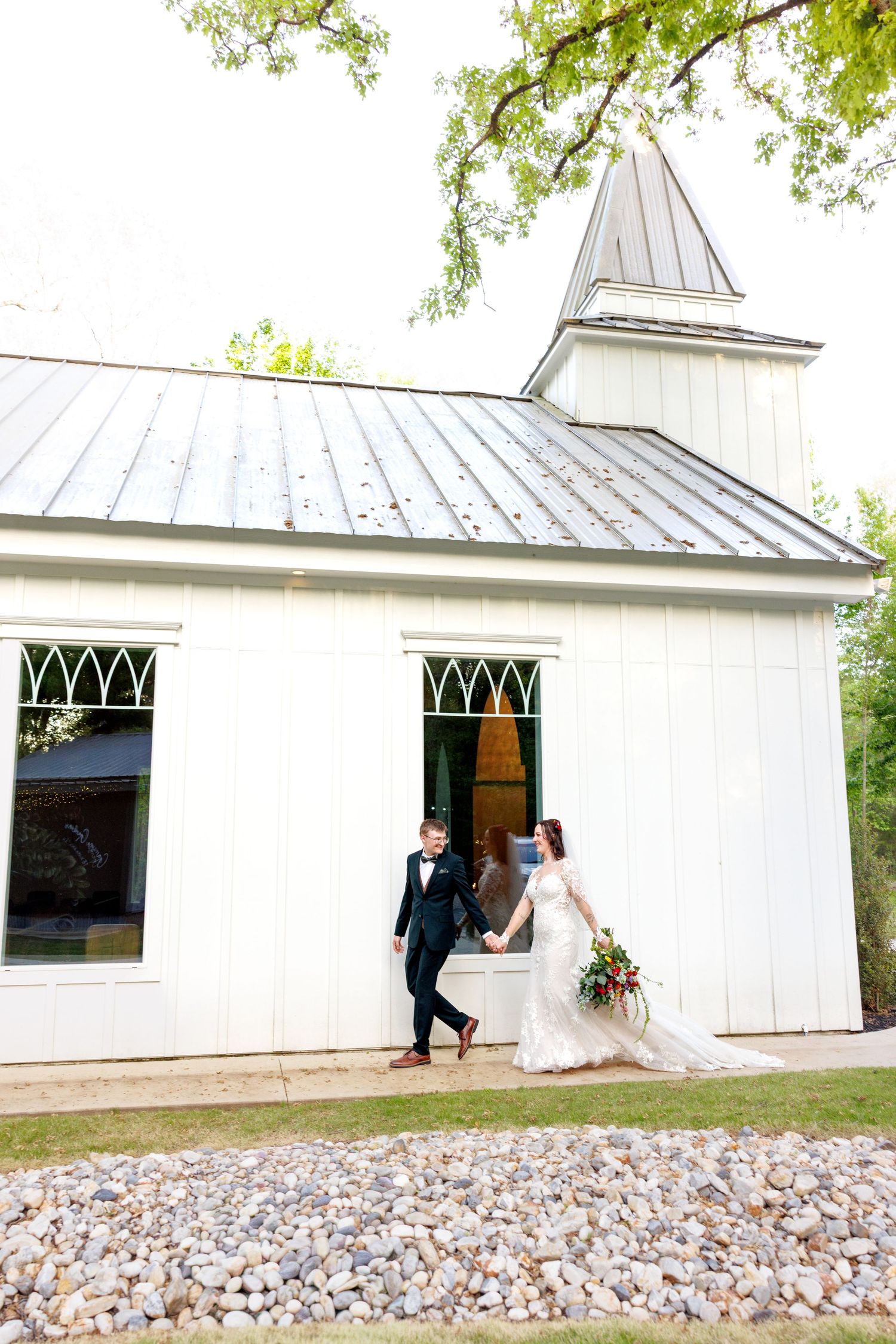 Bride and groom hold hands while exiting a charming white country chapel with a metal steeple roof.