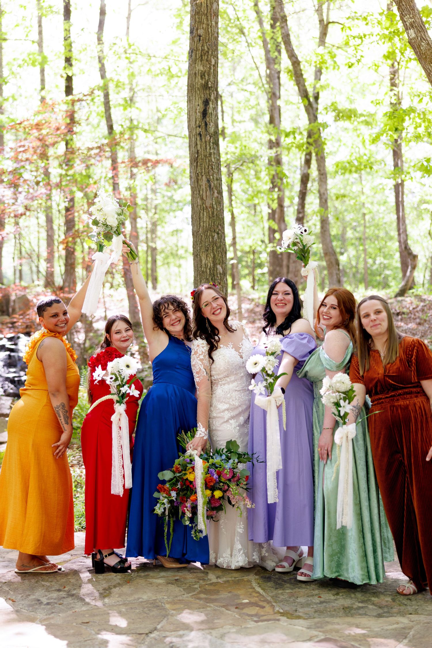 Bride in lace gown poses with six bridesmaids in colorful mismatched dresses holding bouquets in a sunlit forest.