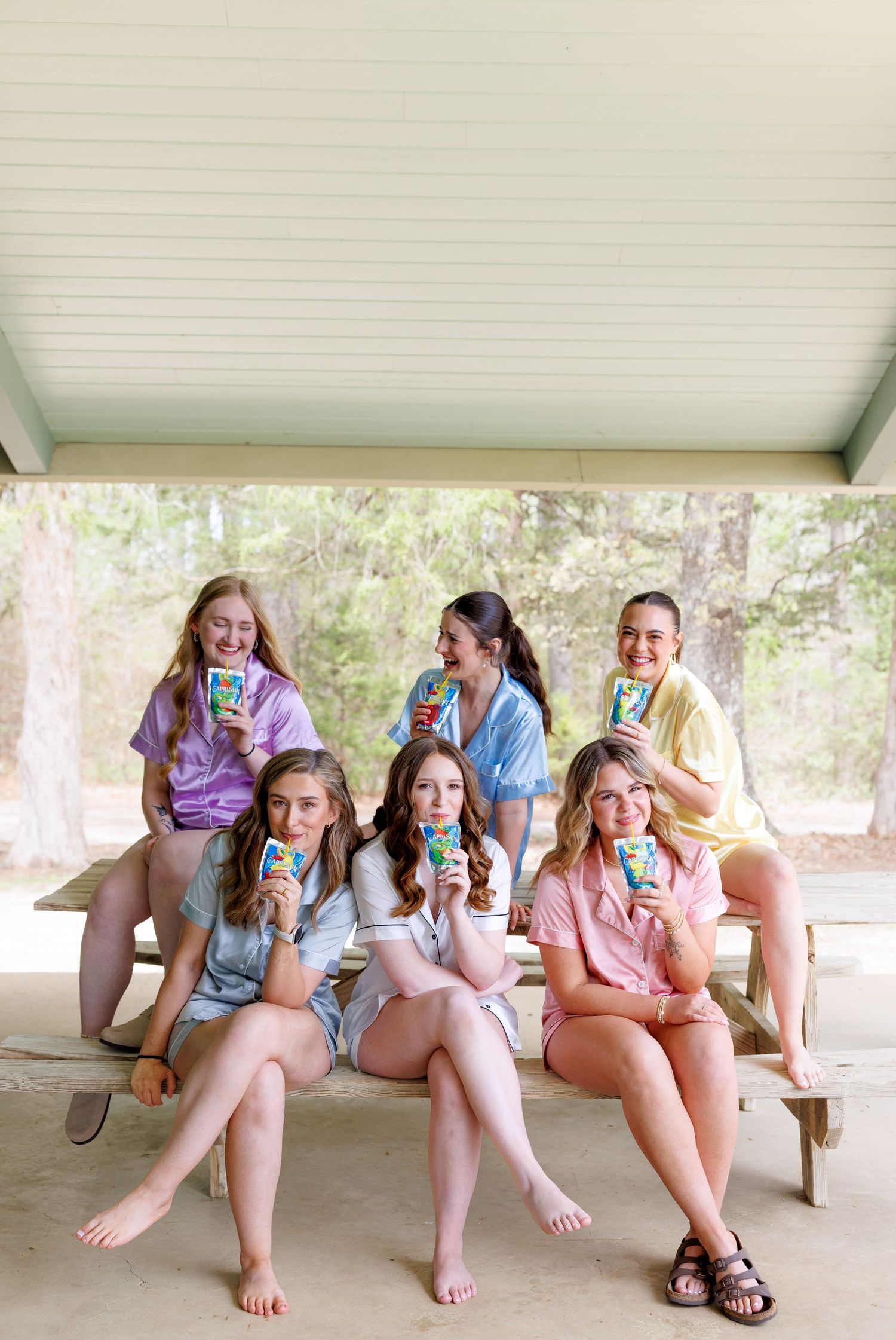 Six young women in colorful matching pajama sets sit at a picnic table outdoors, smiling and holding juice boxes.