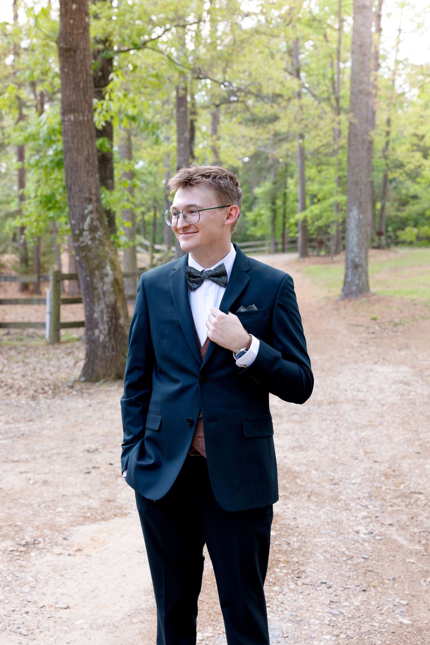 Young man in black tuxedo with bow tie and glasses smiling outdoors on a gravel path surrounded by trees.