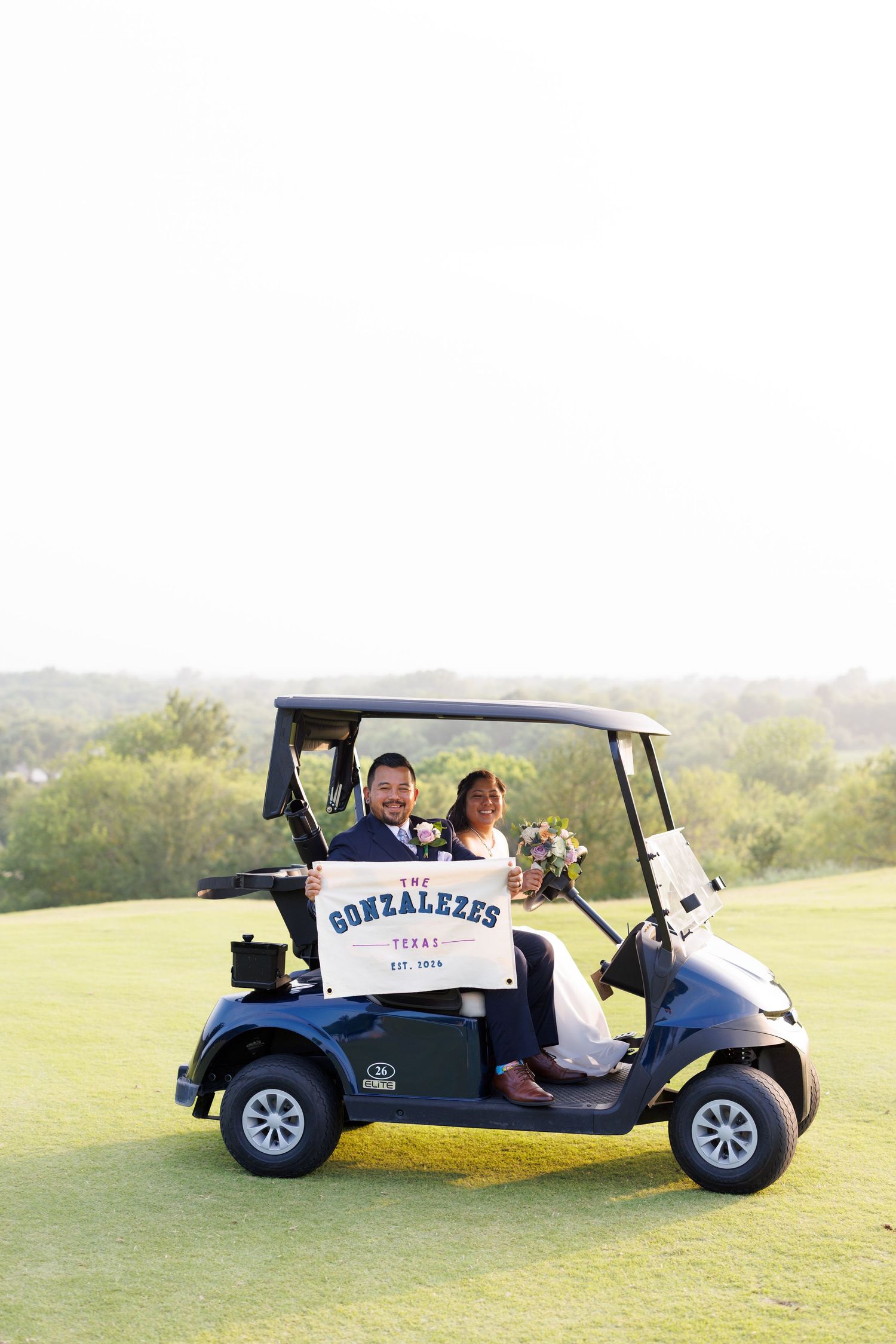 Newlywed couple riding a golf cart holding a The Gonzalezes Texas Est. 2026 wedding sign on a green course.