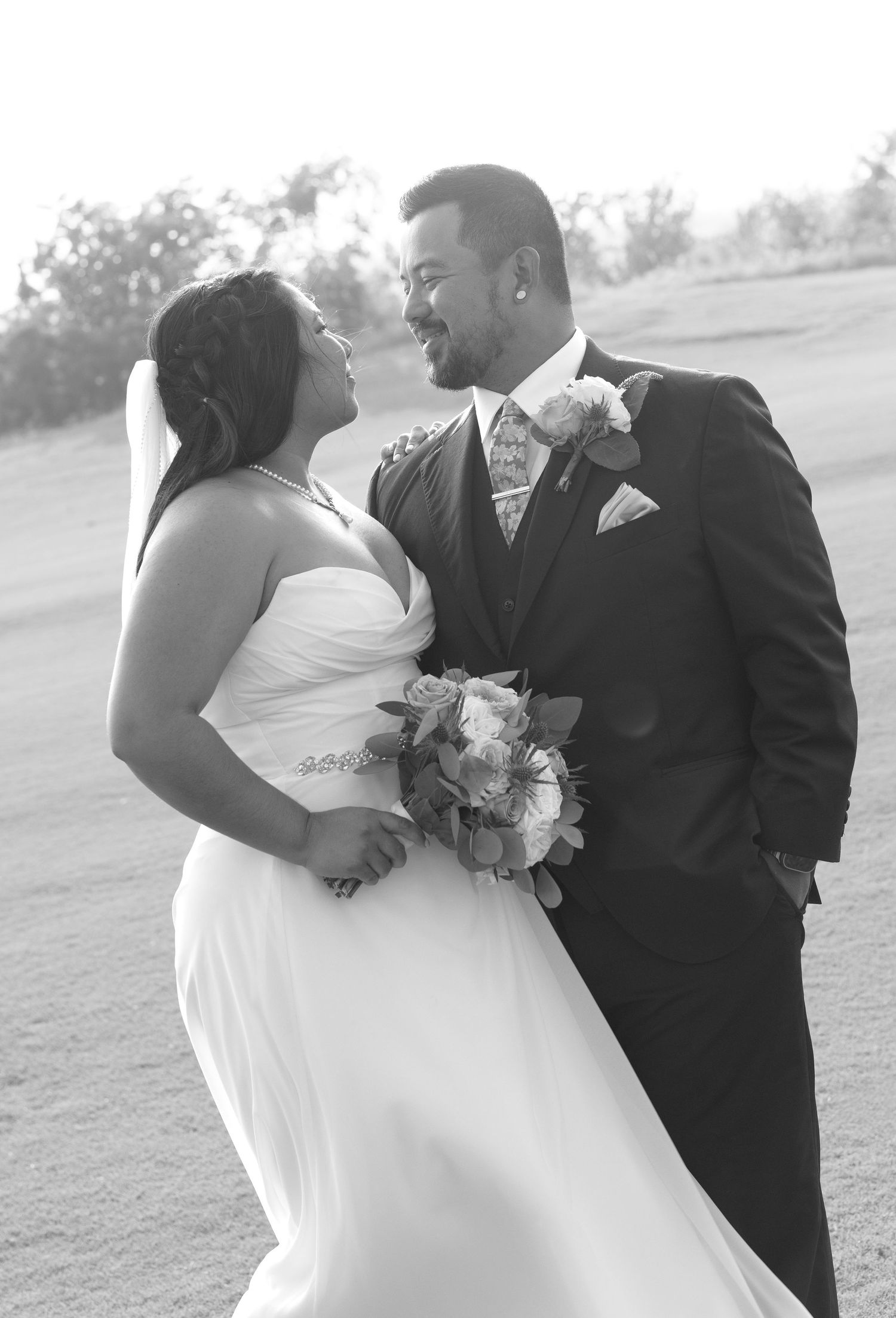 Black and white wedding photo of a smiling bride in white gown holding bouquet and groom in dark suit gazing at each other.