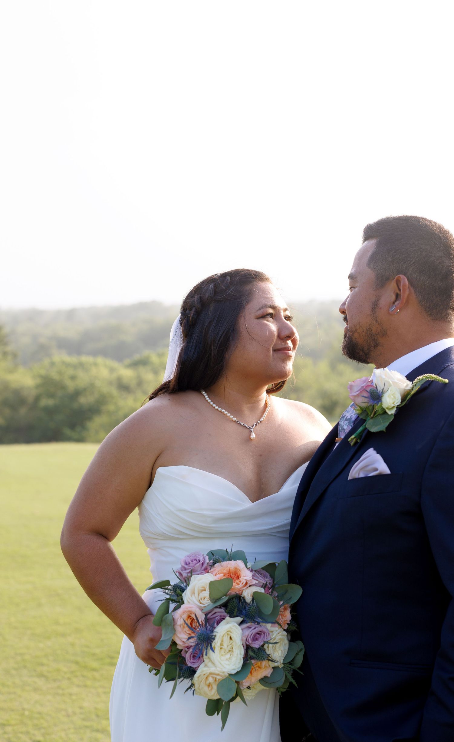 Bride in white gown holding pastel bouquet gazes at groom in navy suit outdoors on lush green golf course.
