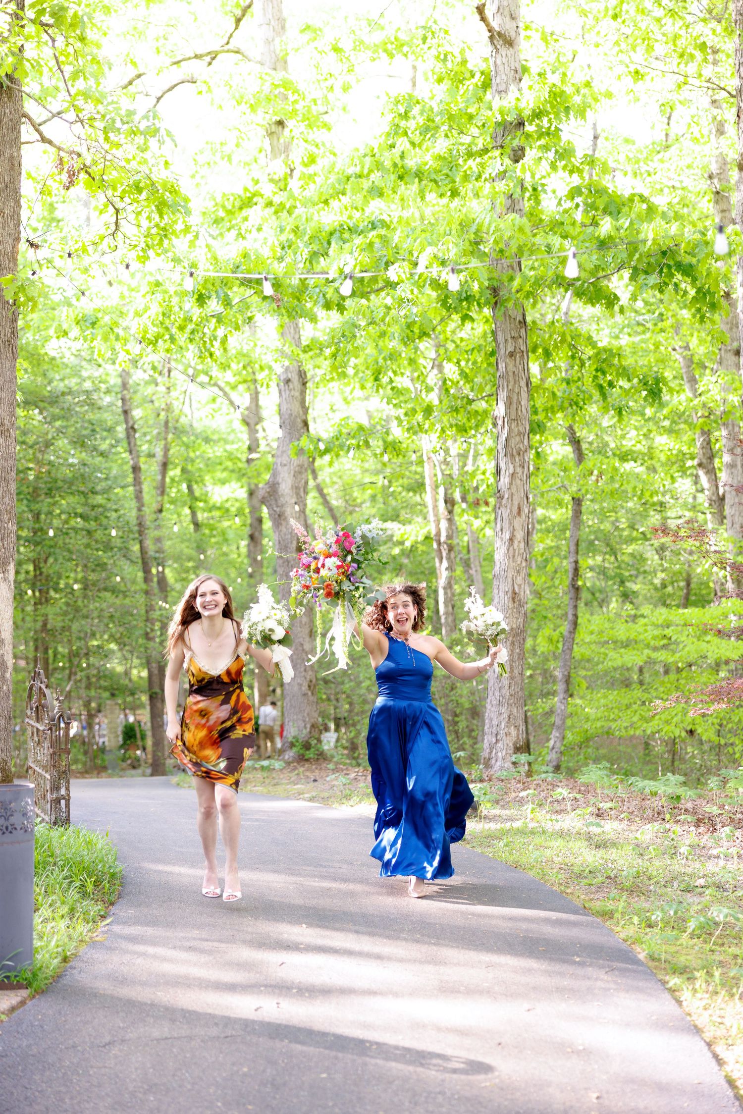 Two women in colorful dresses run joyfully down a tree-lined path holding a vibrant wedding bouquet.