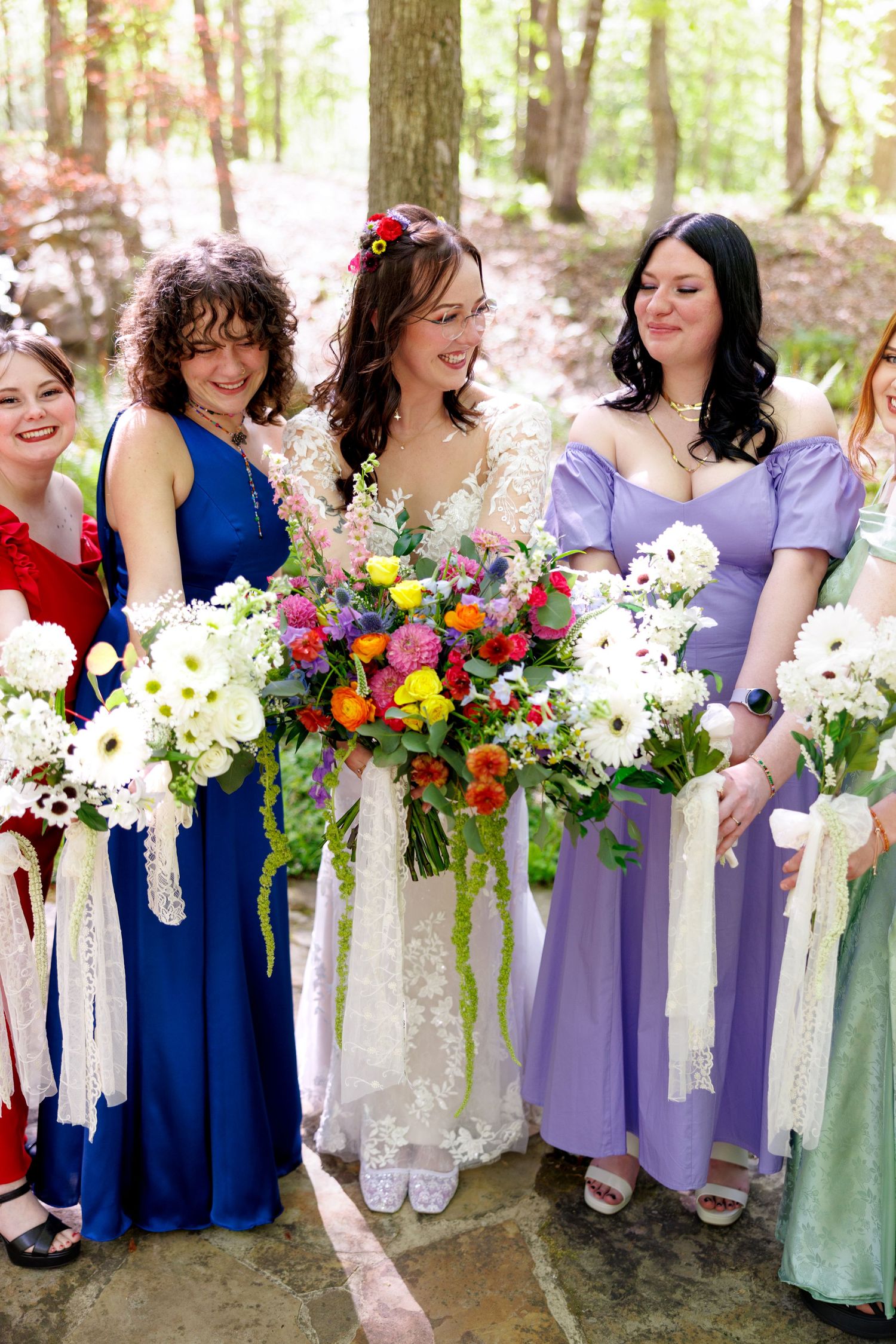 Bride with colorful bouquet poses with bridesmaids in blue and purple dresses in wooded setting.