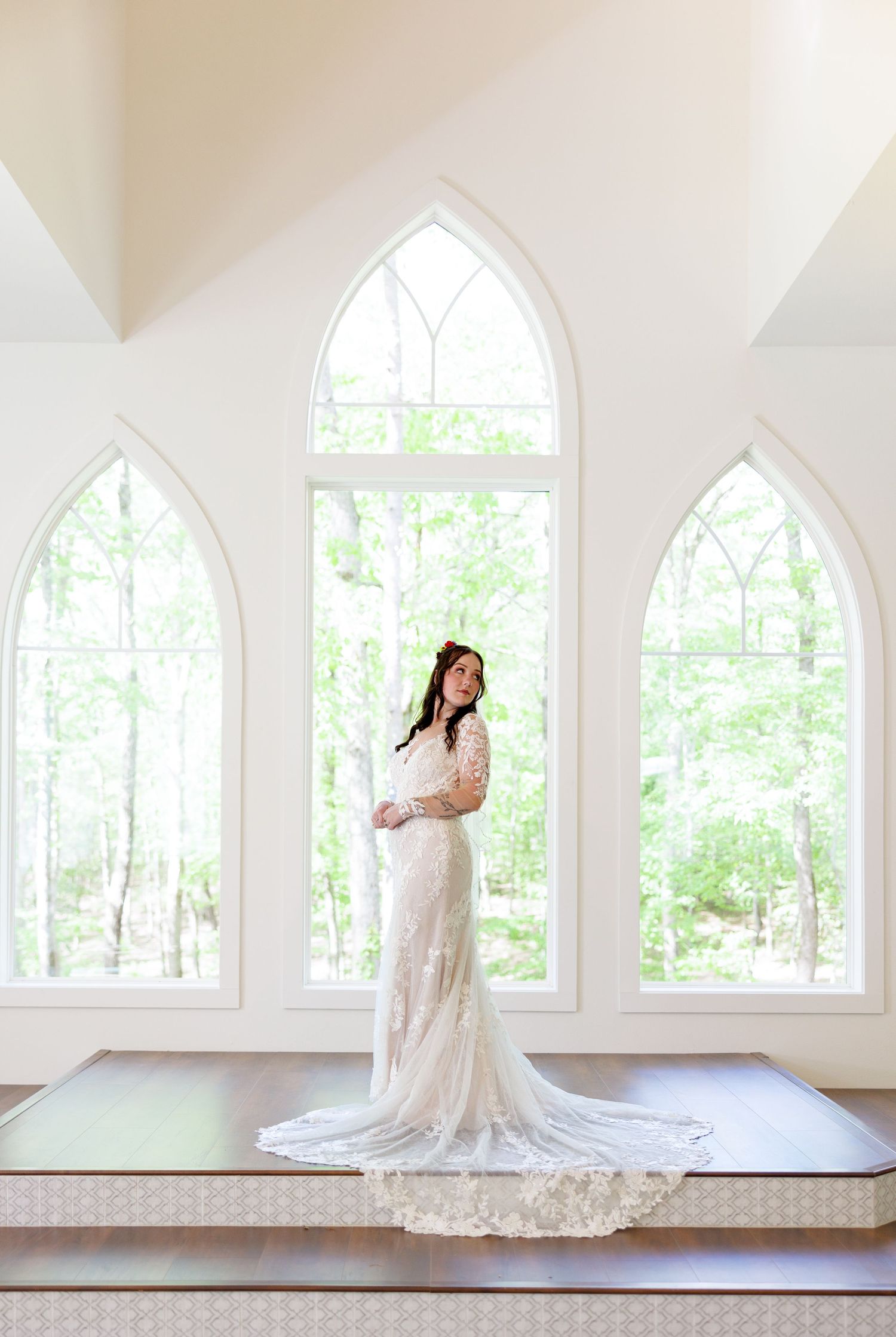 Bride in lace wedding gown with long train standing before Gothic arch windows in a bright white chapel.