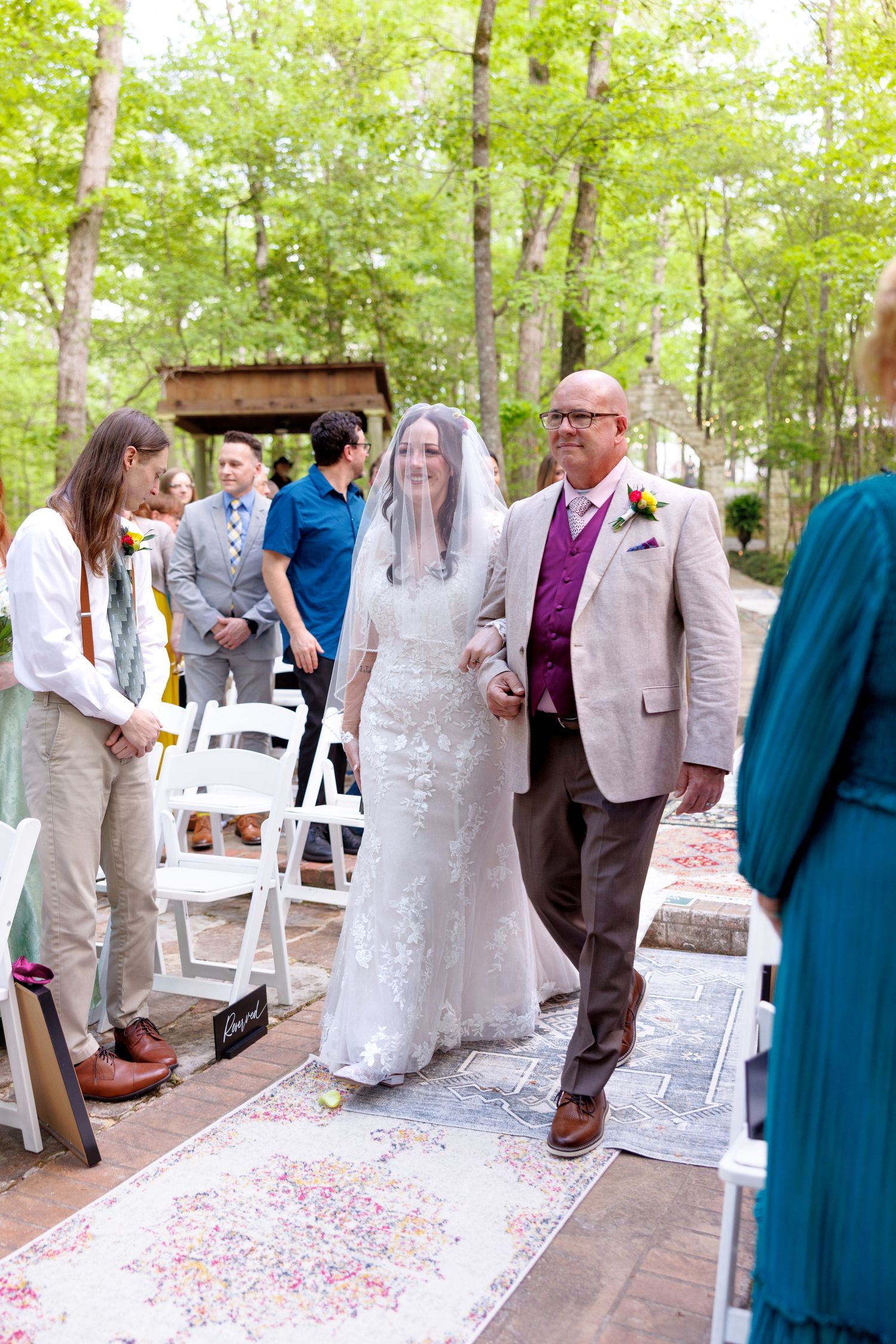 Bride walks down outdoor aisle with escort on colorful rug surrounded by wedding guests and lush green trees.