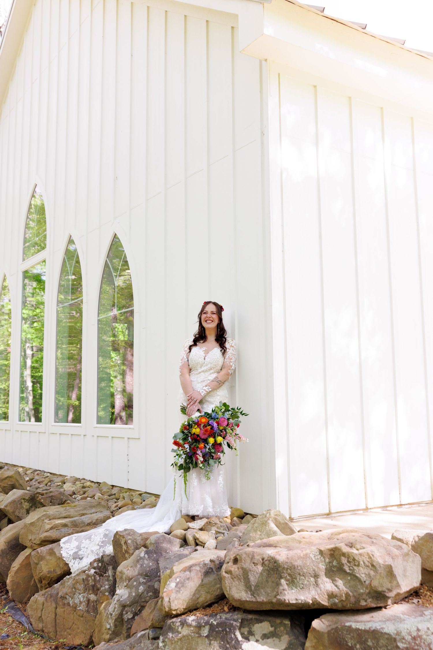 Bride in fitted lace wedding gown holding colorful bouquet standing on rocks beside white chapel exterior.