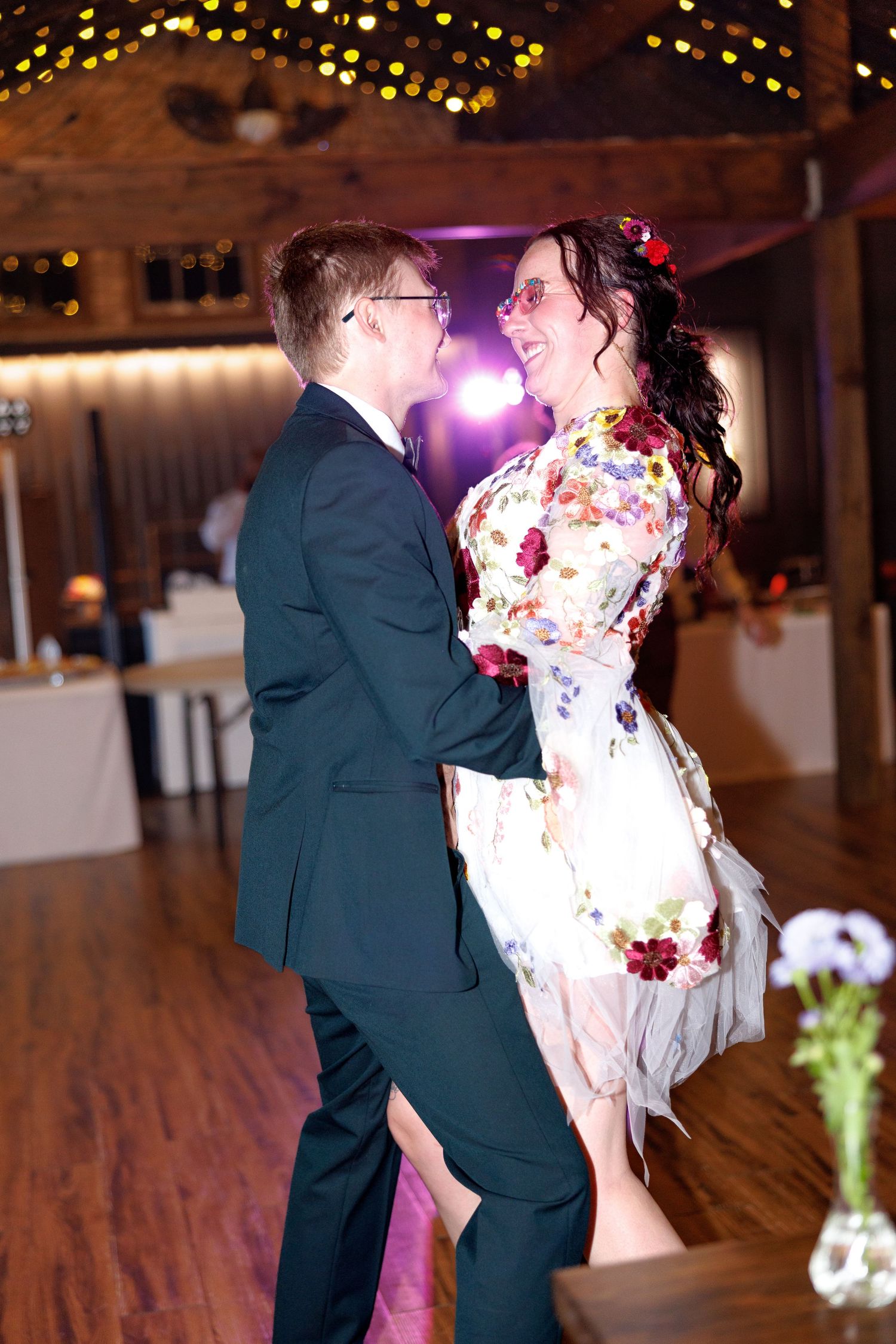 Couple sharing first dance at wedding reception, bride in floral dress with pink sunglasses, groom in dark suit.
