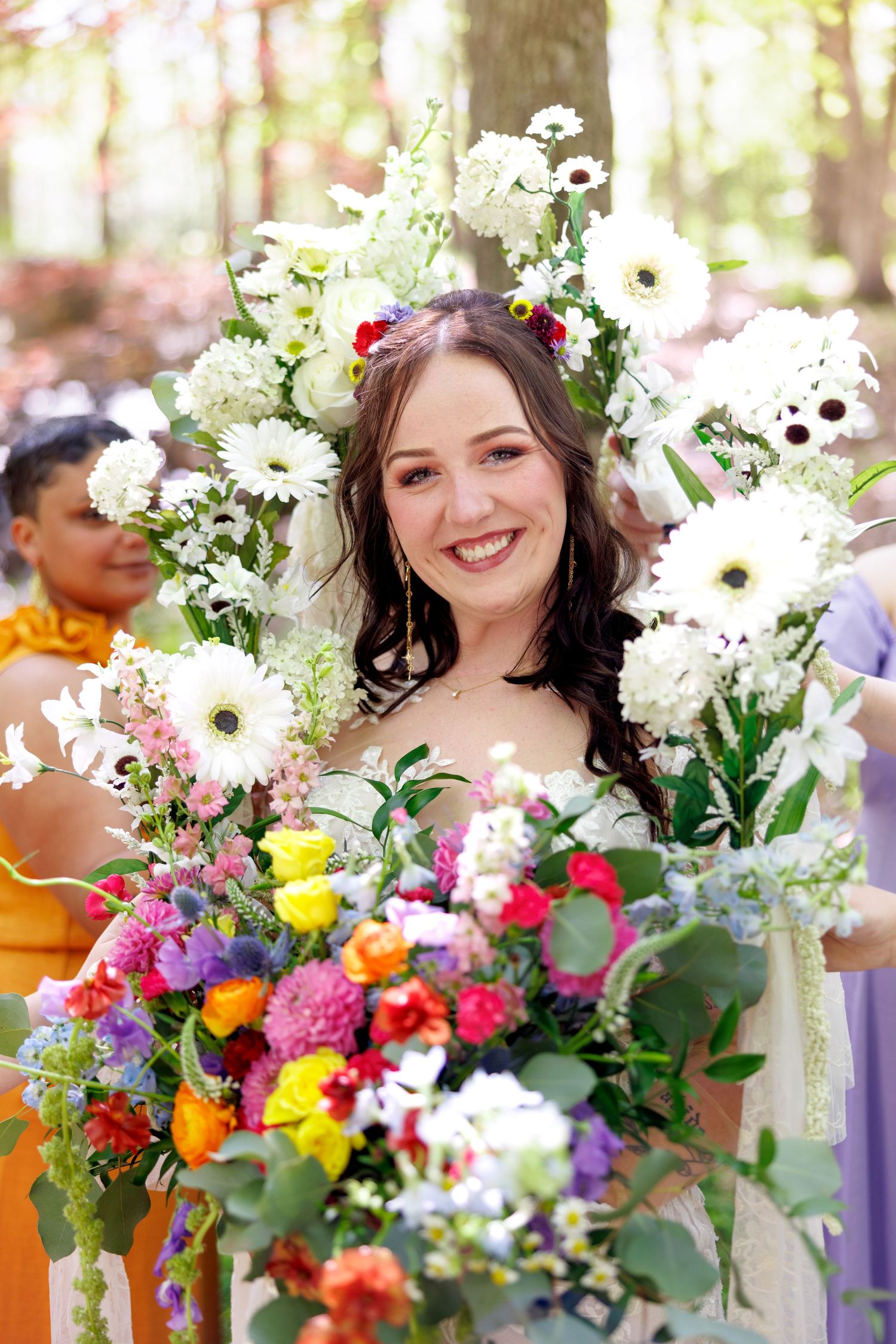 Smiling bride holding colorful bouquet surrounded by white floral arch in a woodland wedding setting.