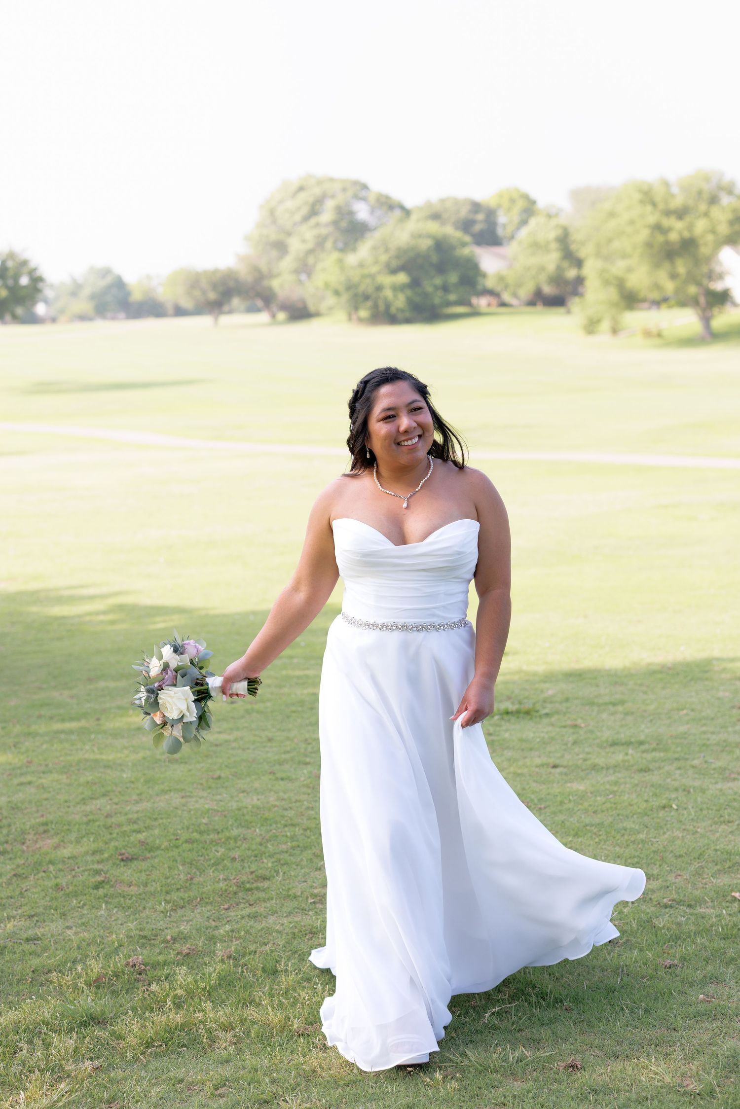 Smiling bride in strapless white gown holding bouquet on lush green golf course lawn on sunny day.