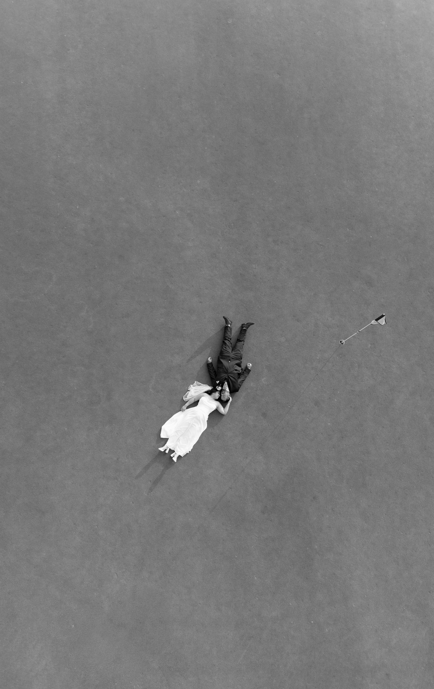 Aerial black and white photo of a bride and groom lying on grass, viewed from above.