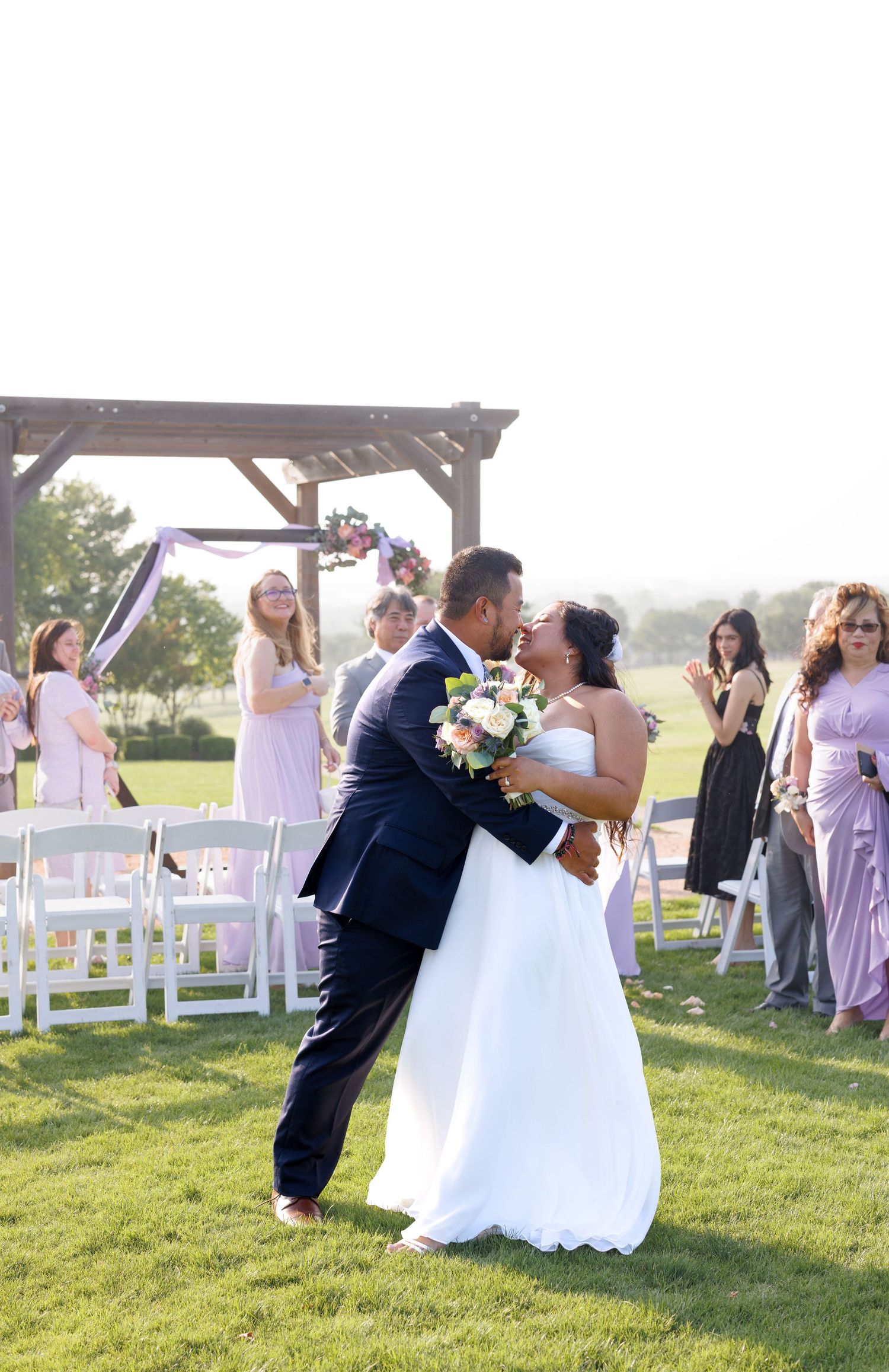 Bride and groom share a romantic kiss on green lawn after outdoor wedding ceremony with guests in lavender dresses.