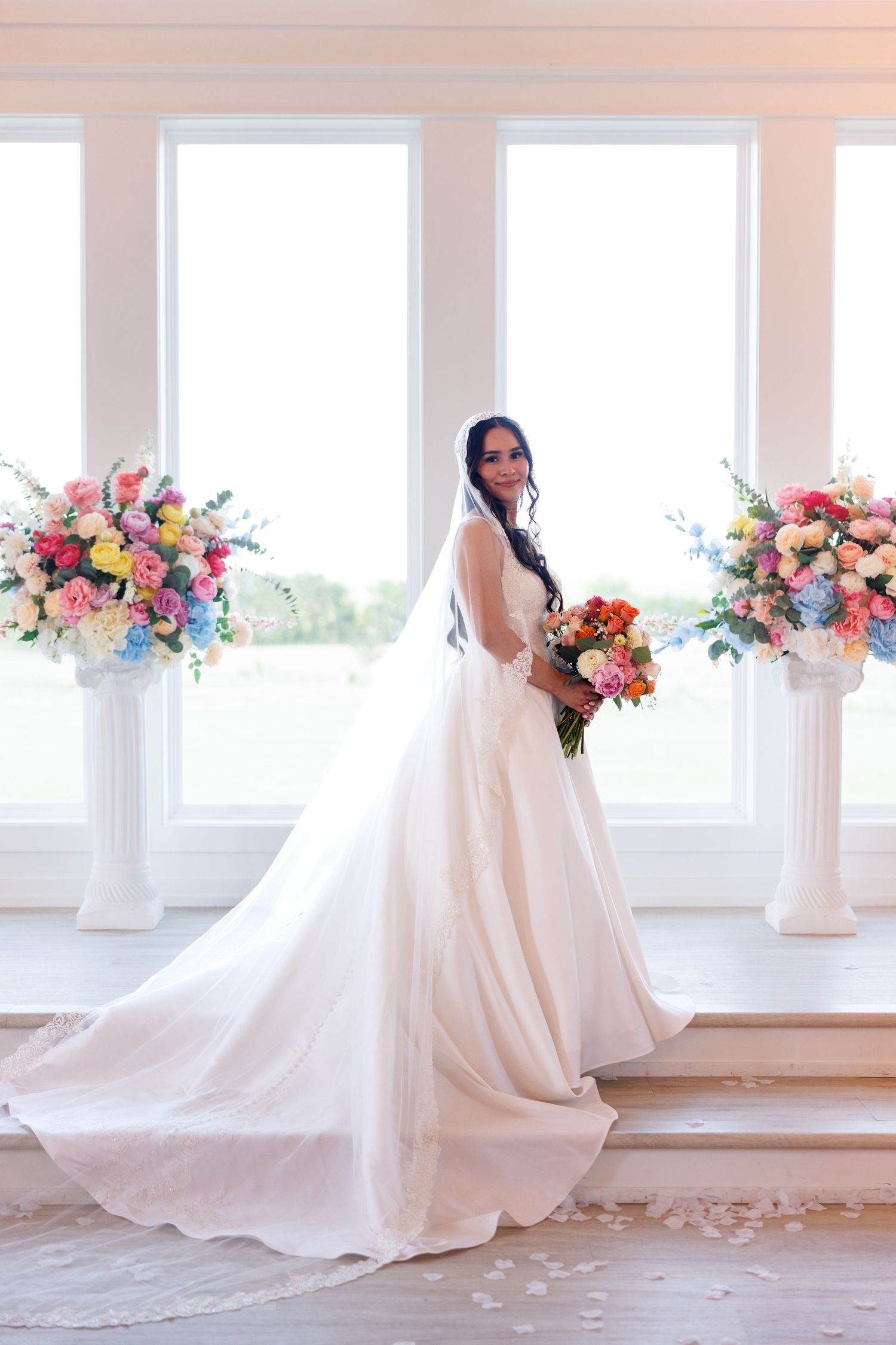 Bride in flowing white gown poses by sunlit windows with pink and orange floral arrangements.