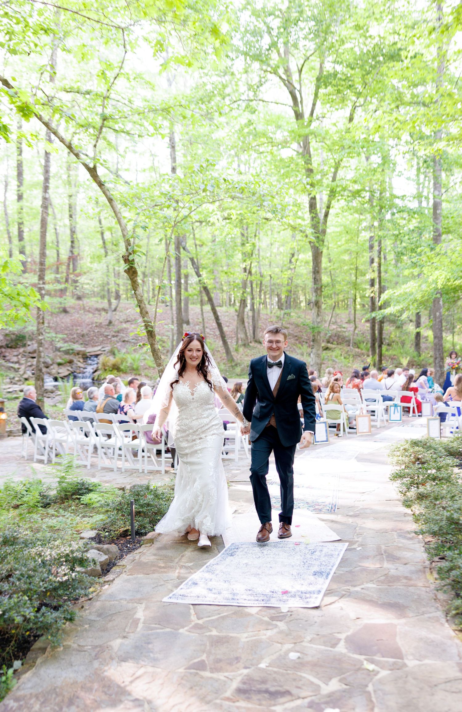 Bride and groom walk hand-in-hand down an outdoor stone aisle surrounded by lush green trees after their wedding ceremony.