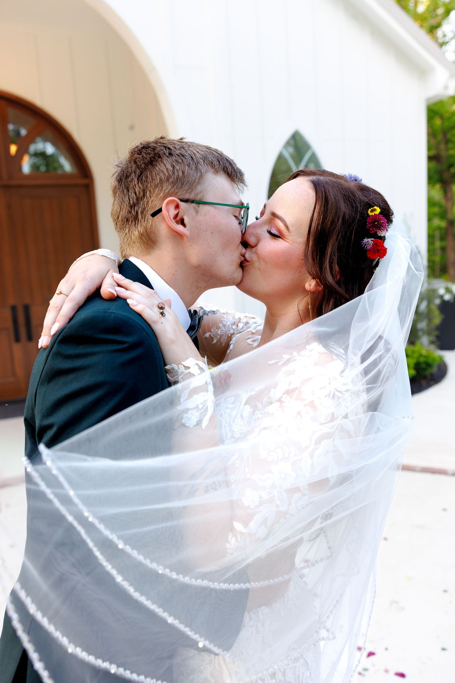 Newlyweds sharing a romantic kiss under a flowing white veil outside a white chapel after their wedding ceremony.