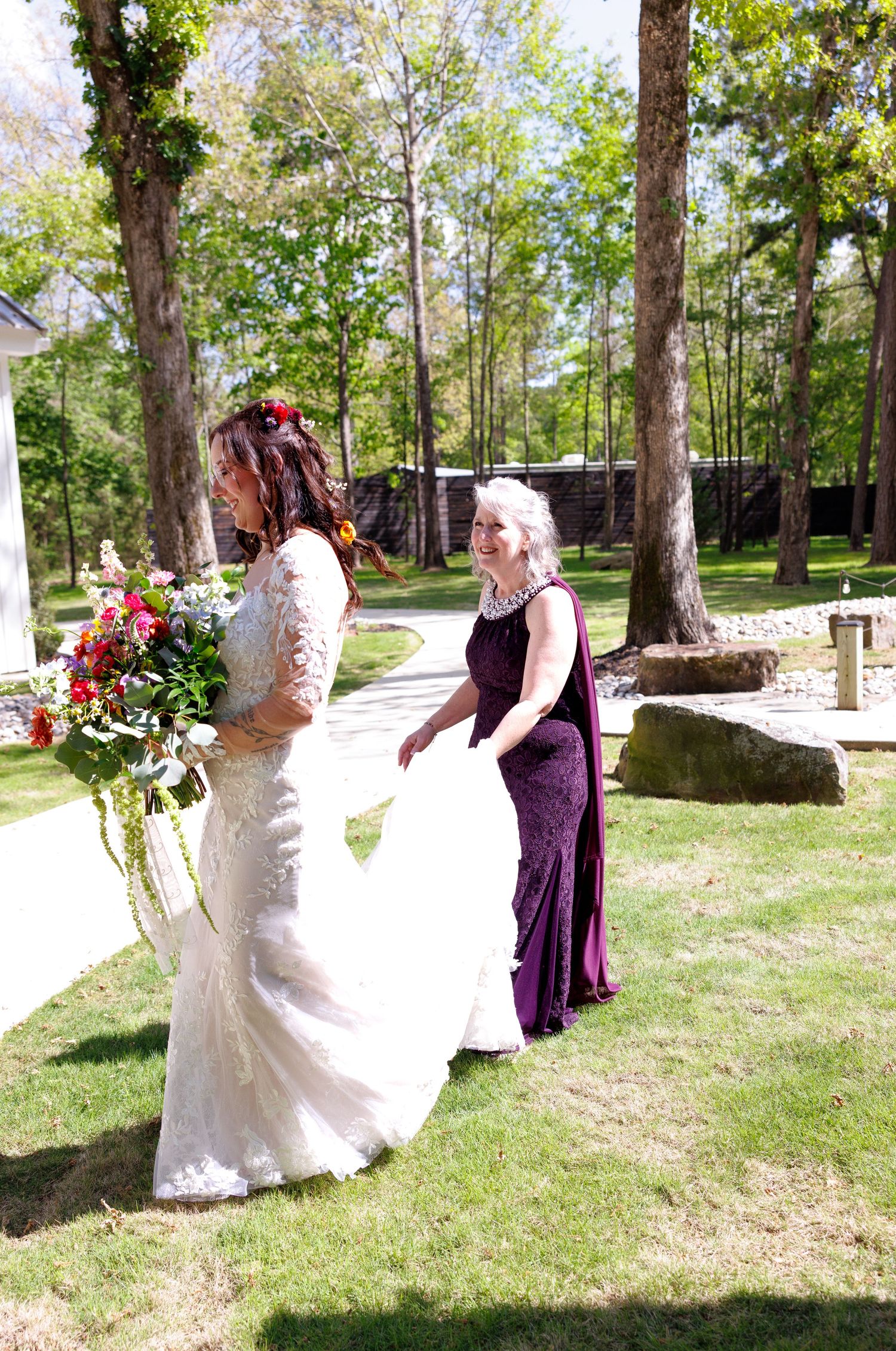 Bride in white lace gown holding bouquet walks outdoors with older woman in purple dress carrying her train.