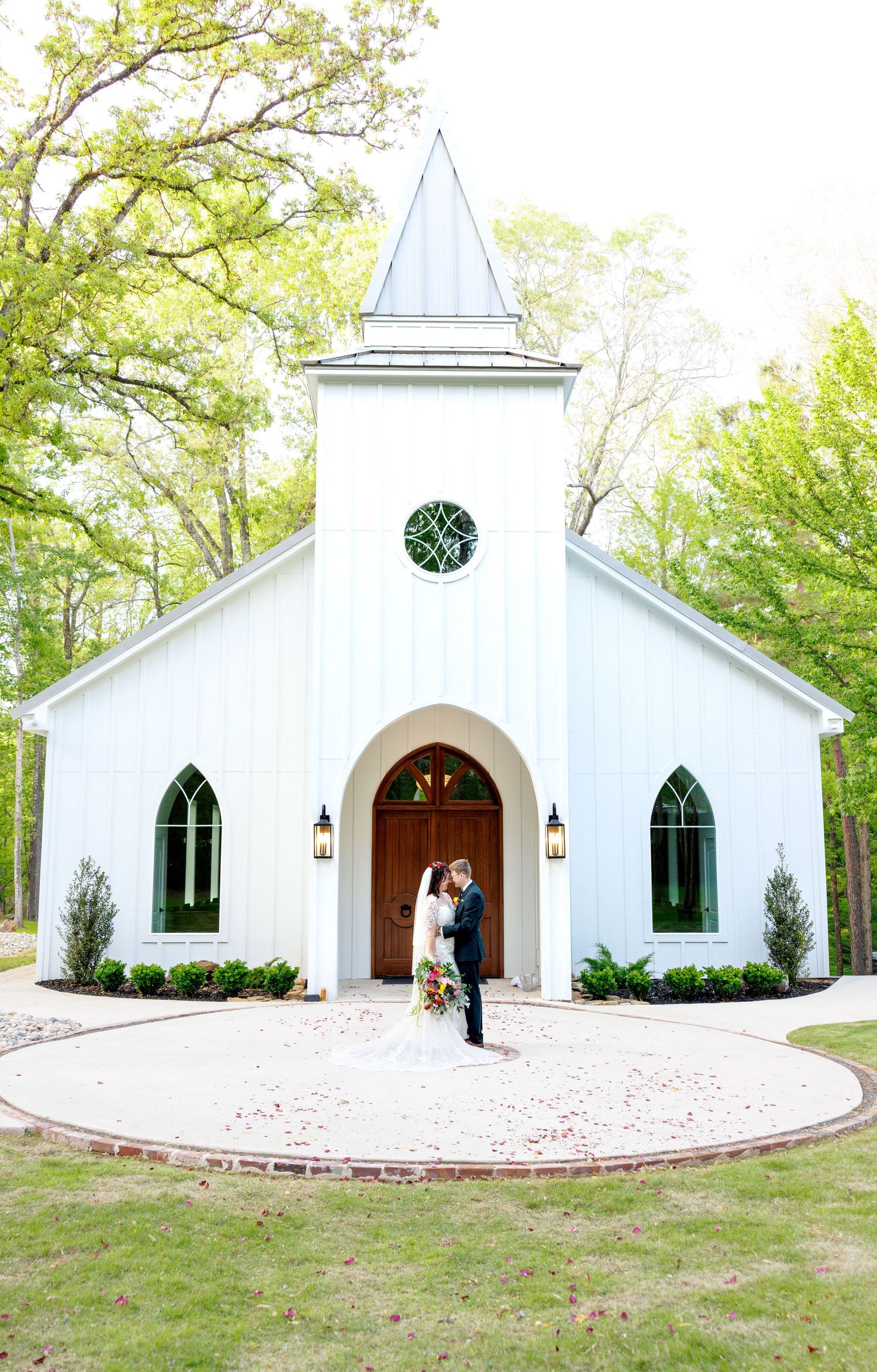 Newlywed couple embraces in front of a charming white wedding chapel with a steeple surrounded by spring trees.