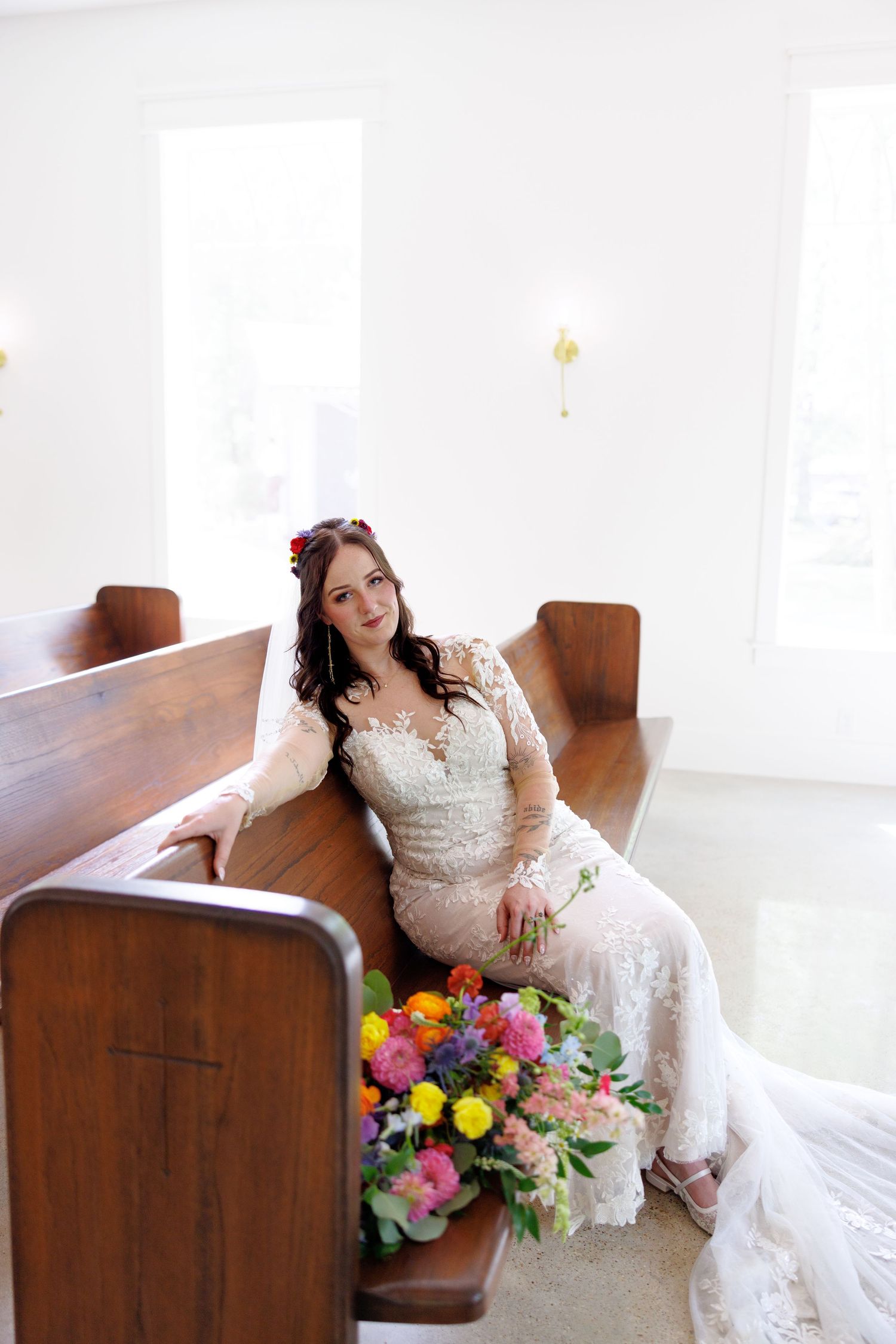 Bride in lace gown with floral crown sits on wooden church pew holding colorful bouquet in bright white chapel.