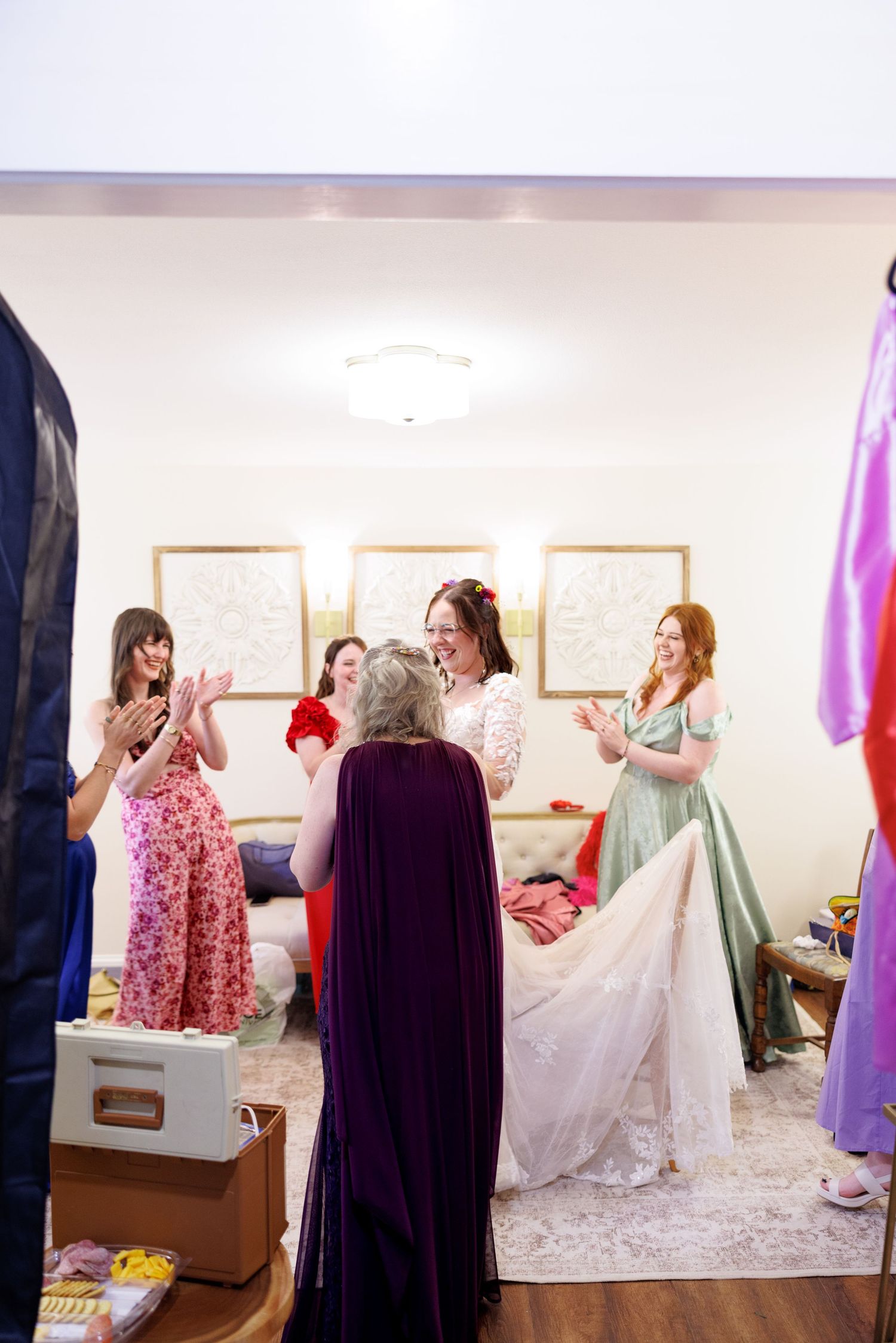 Bride in white lace gown celebrated by bridesmaids in colorful dresses inside a bright dressing room.