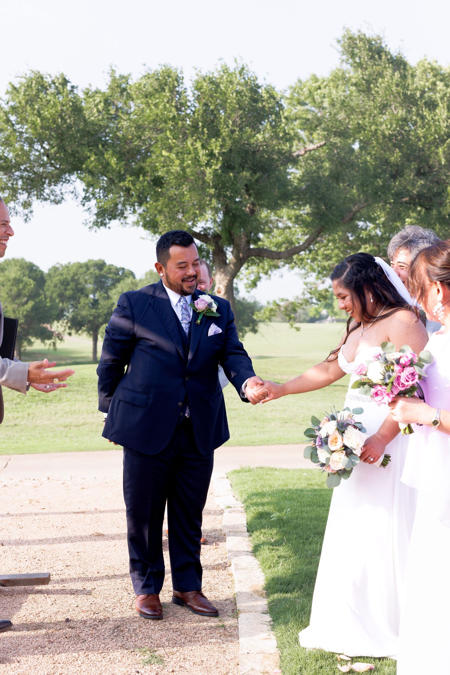 Smiling groom in navy suit reaches for brides hand during outdoor wedding ceremony on sunny day.