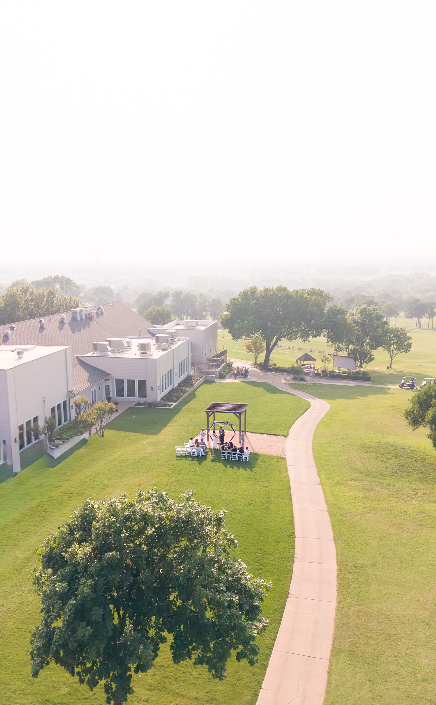 Aerial drone view of an outdoor wedding ceremony on lush green golf course grounds beside a modern venue building.