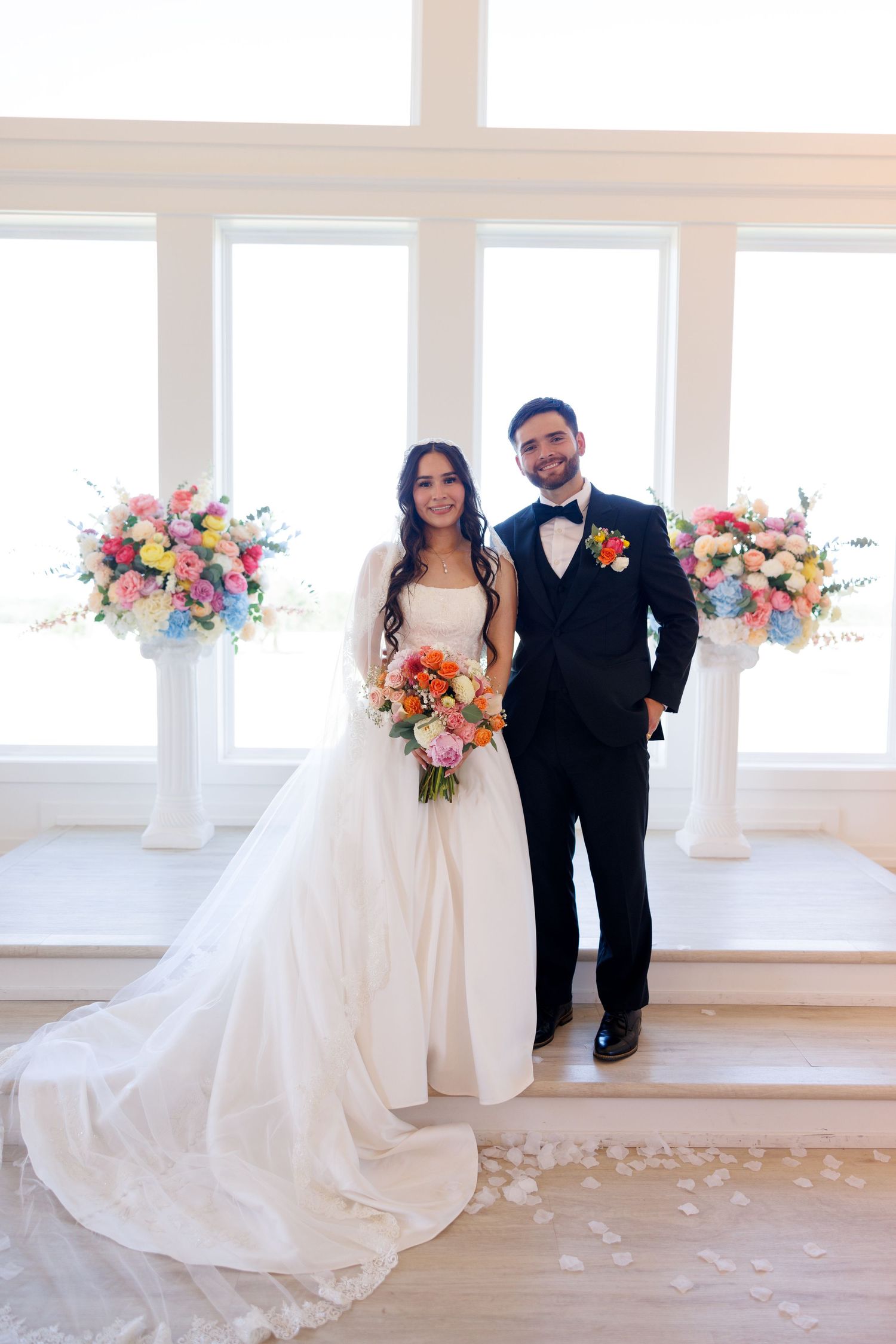 Bride and groom share intimate moments in sunlit white room with pink and blue floral decorations.