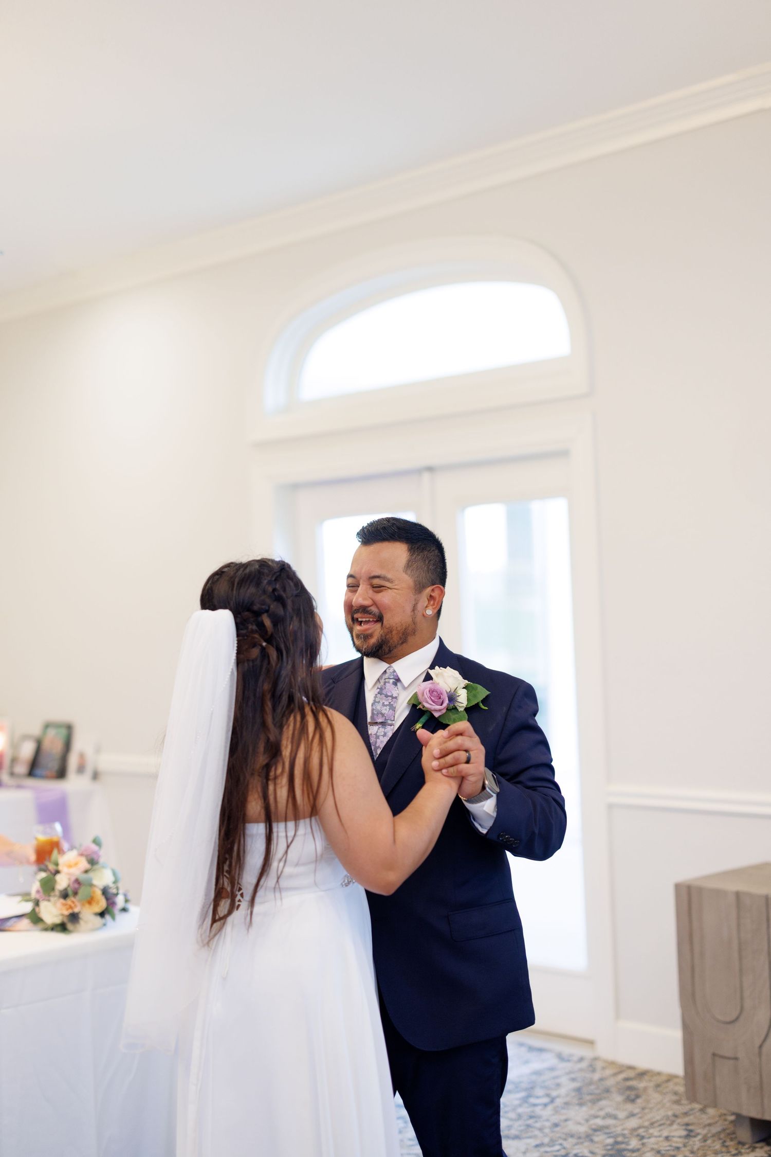 A bride and groom share their first dance, smiling joyfully in an elegant white reception hall.