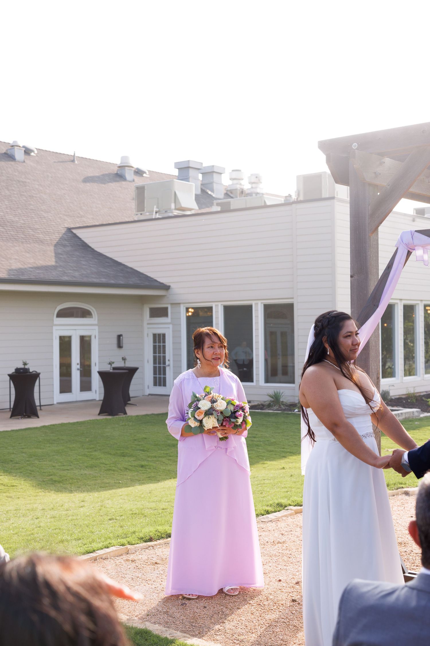 Bride in white gown stands with bridesmaid in pink dress holding bouquet during outdoor wedding ceremony.