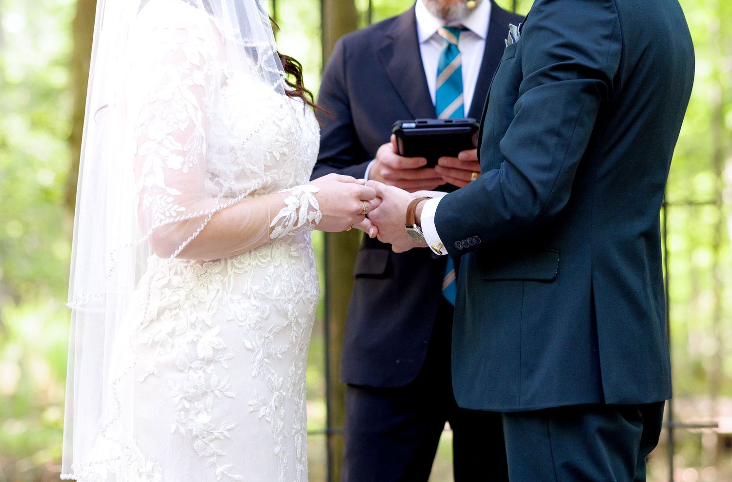 Bride and groom exchanging vows outdoors with officiant holding book during elegant forest wedding ceremony.