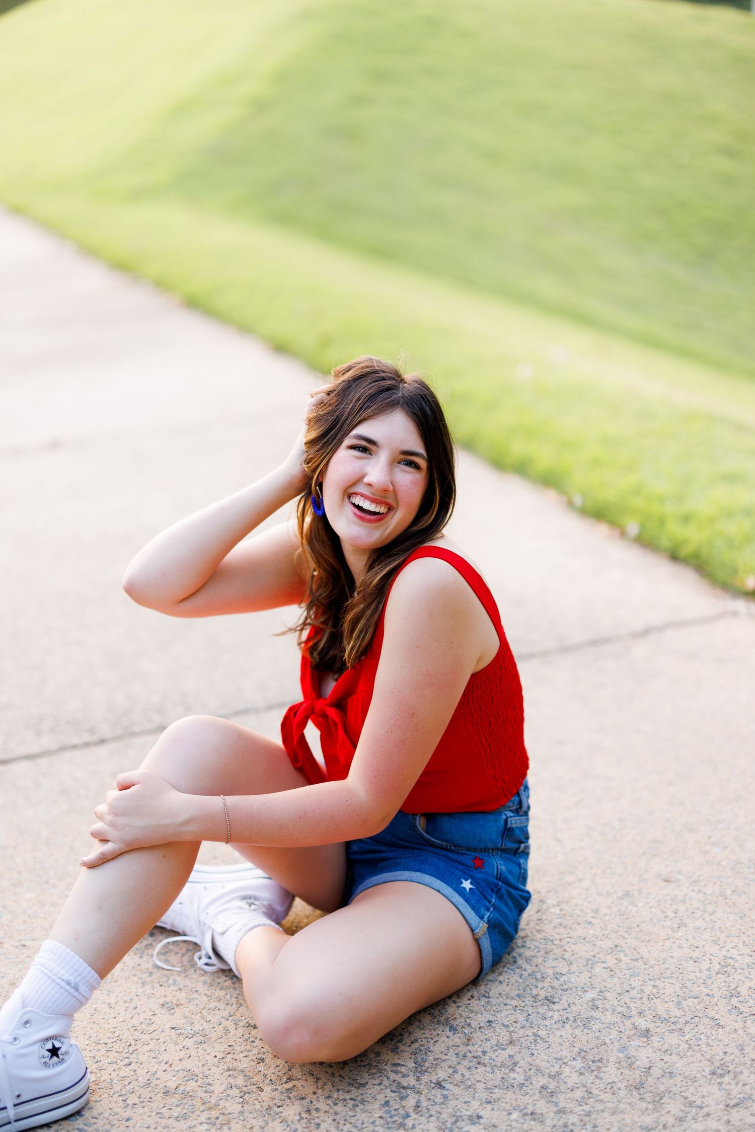 Person in red top and denim shorts sitting playfully on concrete path outdoors.