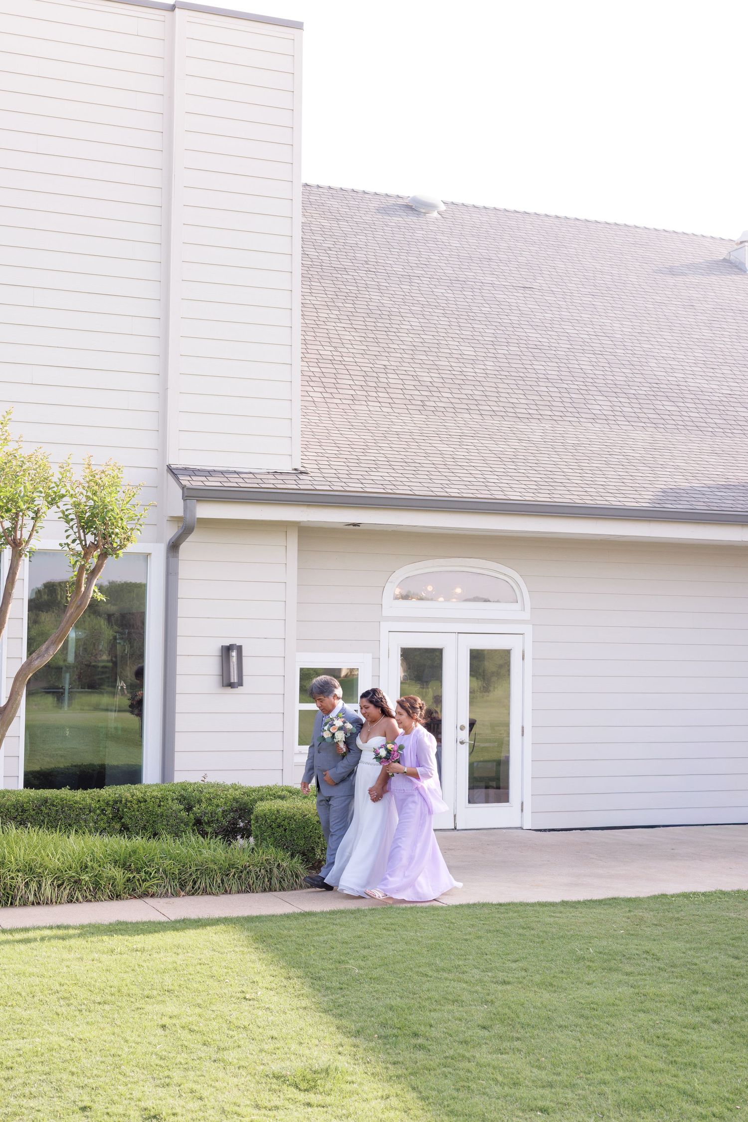 Bride and groom with wedding party posing outside elegant white venue with French doors and manicured hedges.