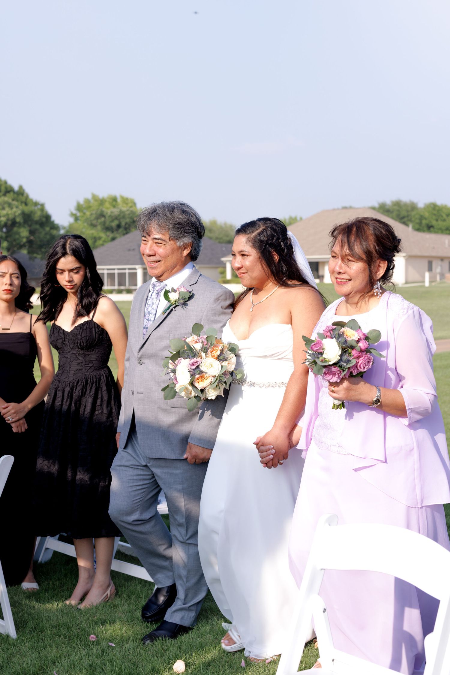 Bride walks down outdoor aisle with parents, holding floral bouquet, bridesmaids in black dresses behind her.