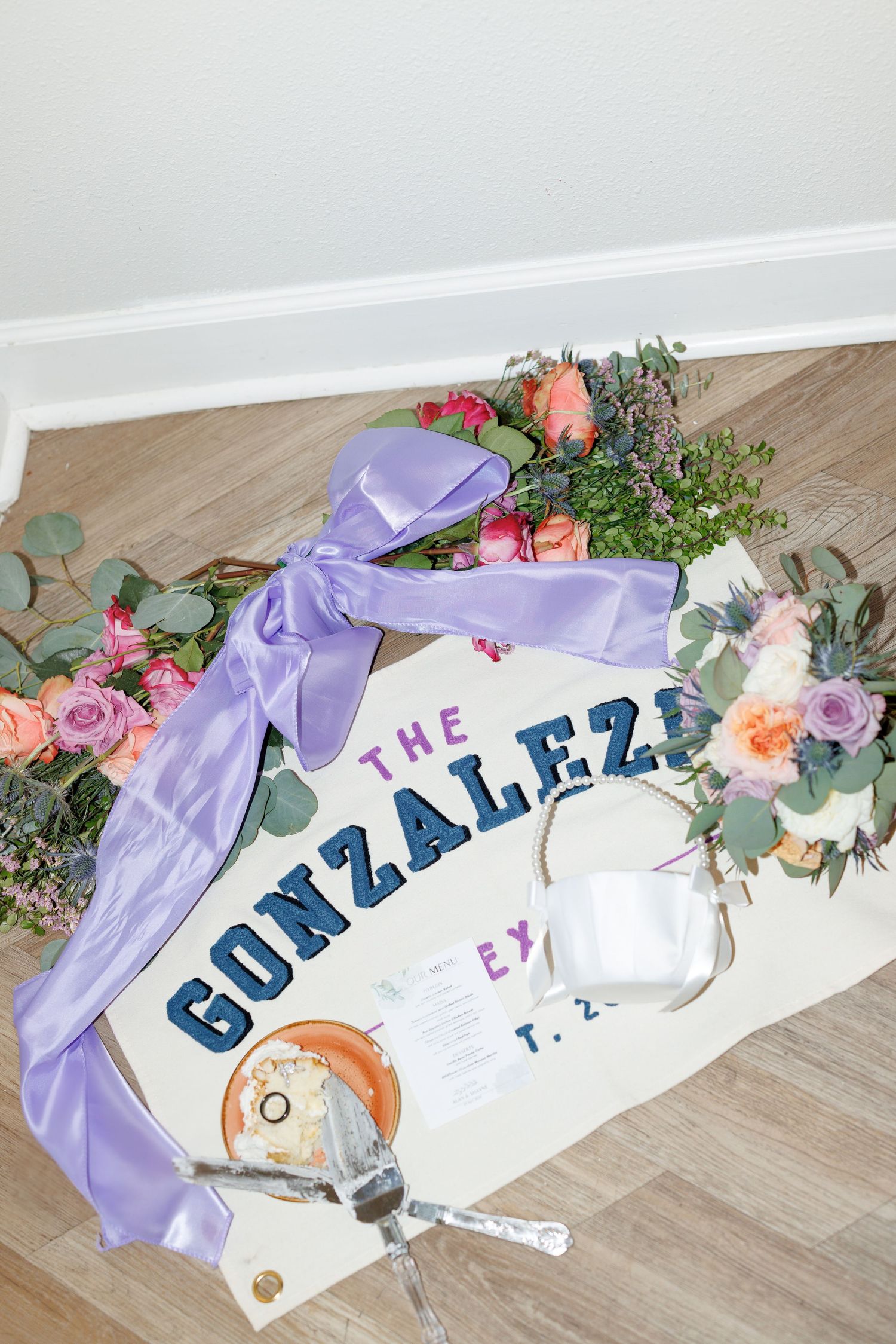 Wedding flat lay featuring The Gonzalez sign with purple ribbon, floral bouquets, rings, and invitation on wood floor.