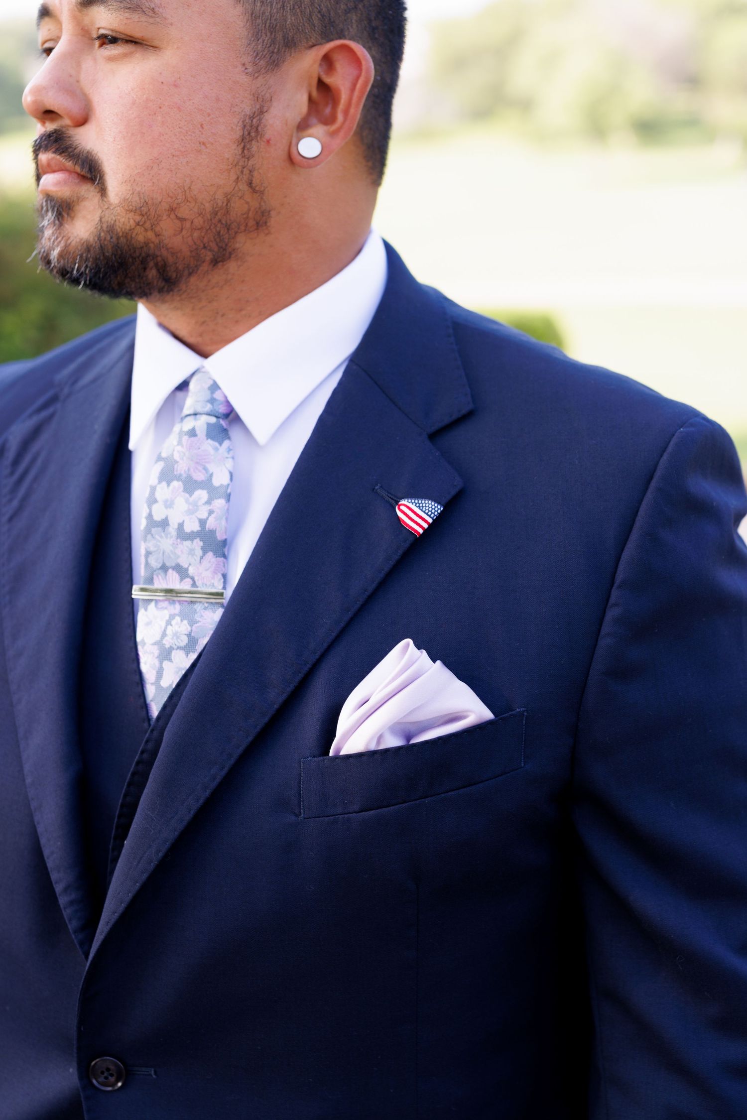 Man in navy suit with floral tie, pocket square, lapel pin, and pearl earring outdoors.