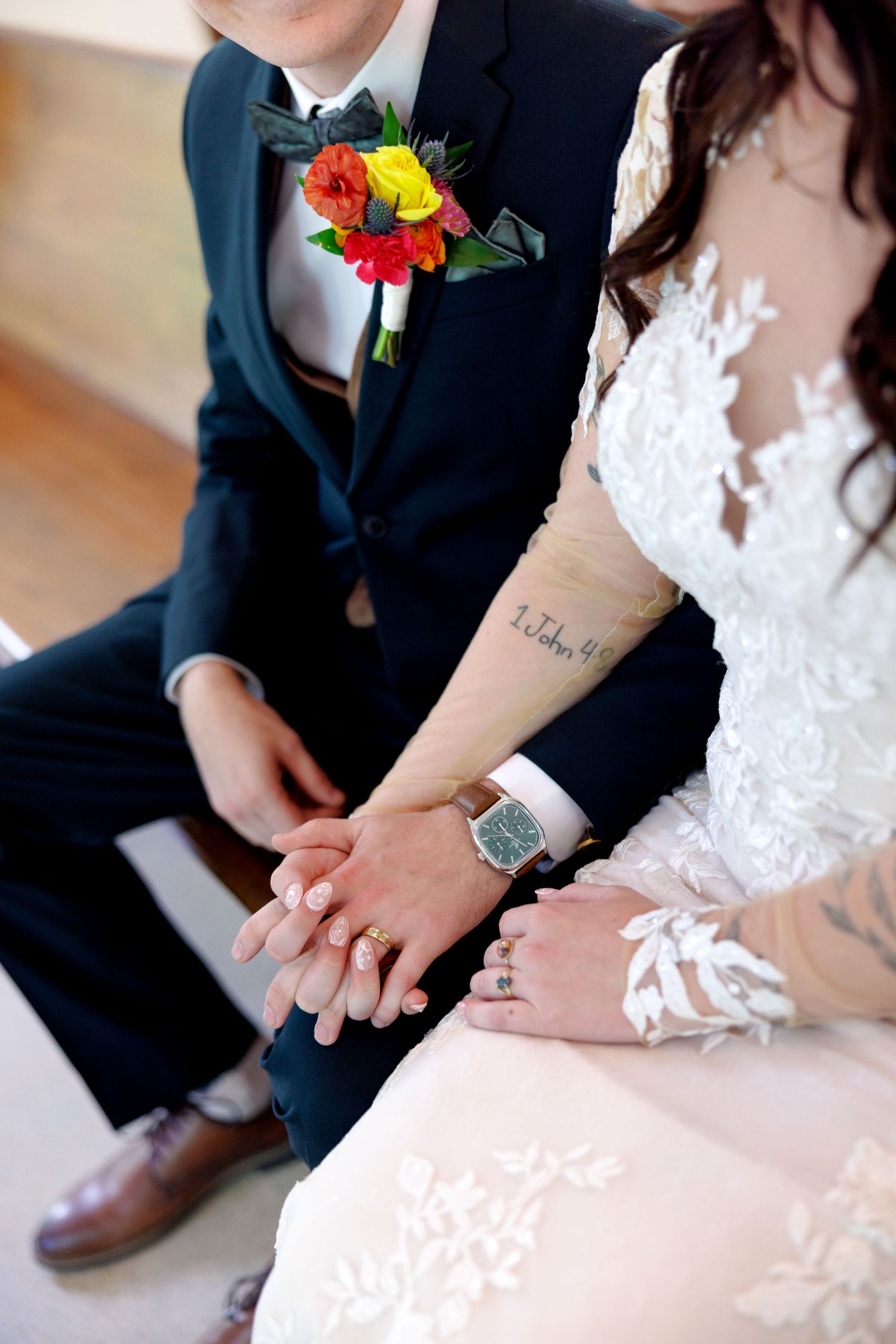 Bride and groom holding hands showing wedding rings, with colorful boutonniere and lace wedding dress details.