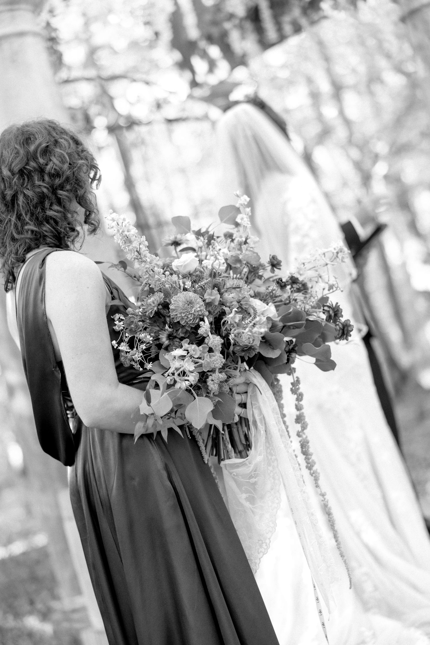 Bridesmaid in elegant gown holds large floral bouquet as bride and groom stand at outdoor wedding ceremony.