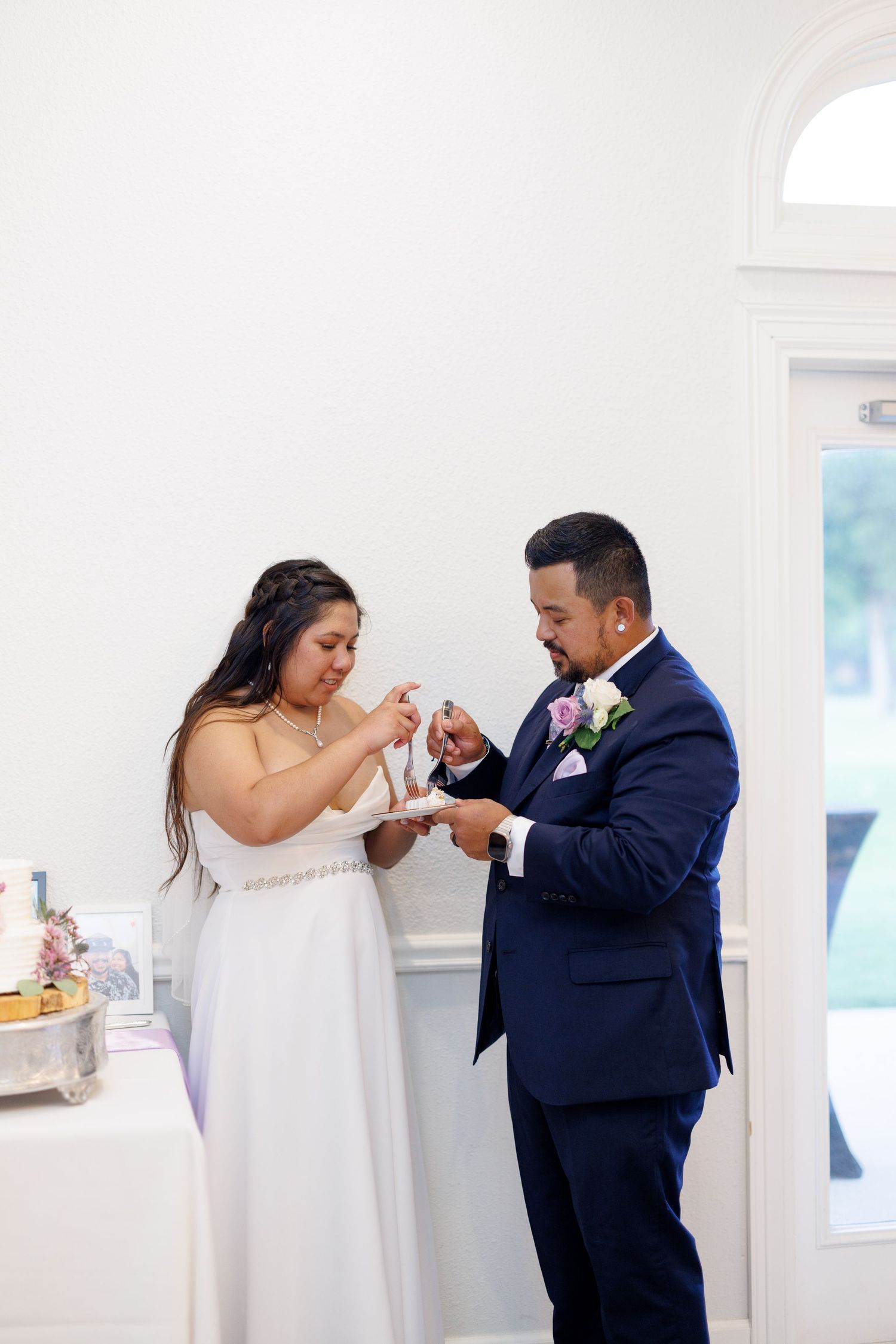Bride and groom cutting their wedding cake together in an elegant white venue, groom wearing a navy suit.