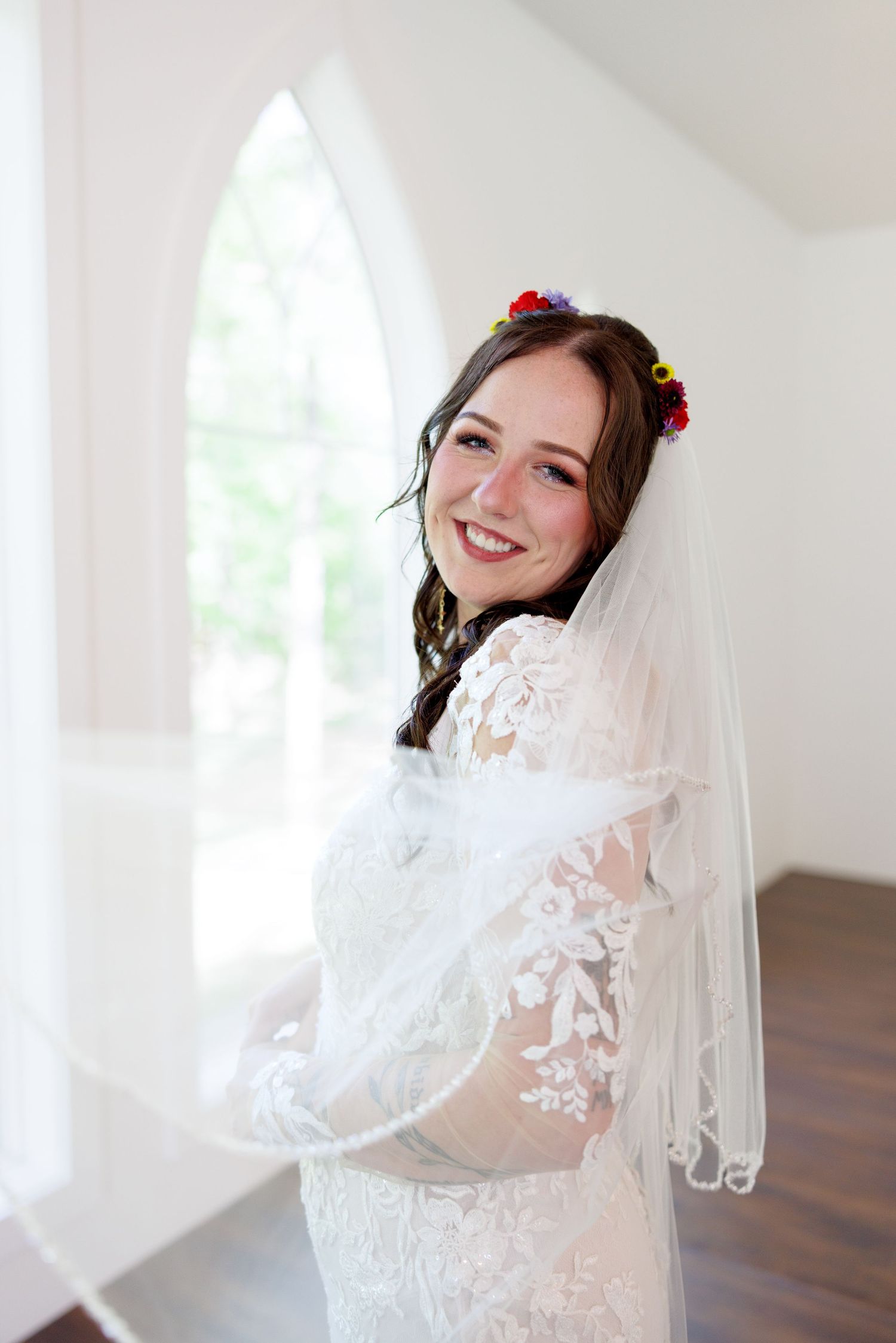 Smiling bride in lace wedding dress and colorful flower crown with white veil, posing by arched window.