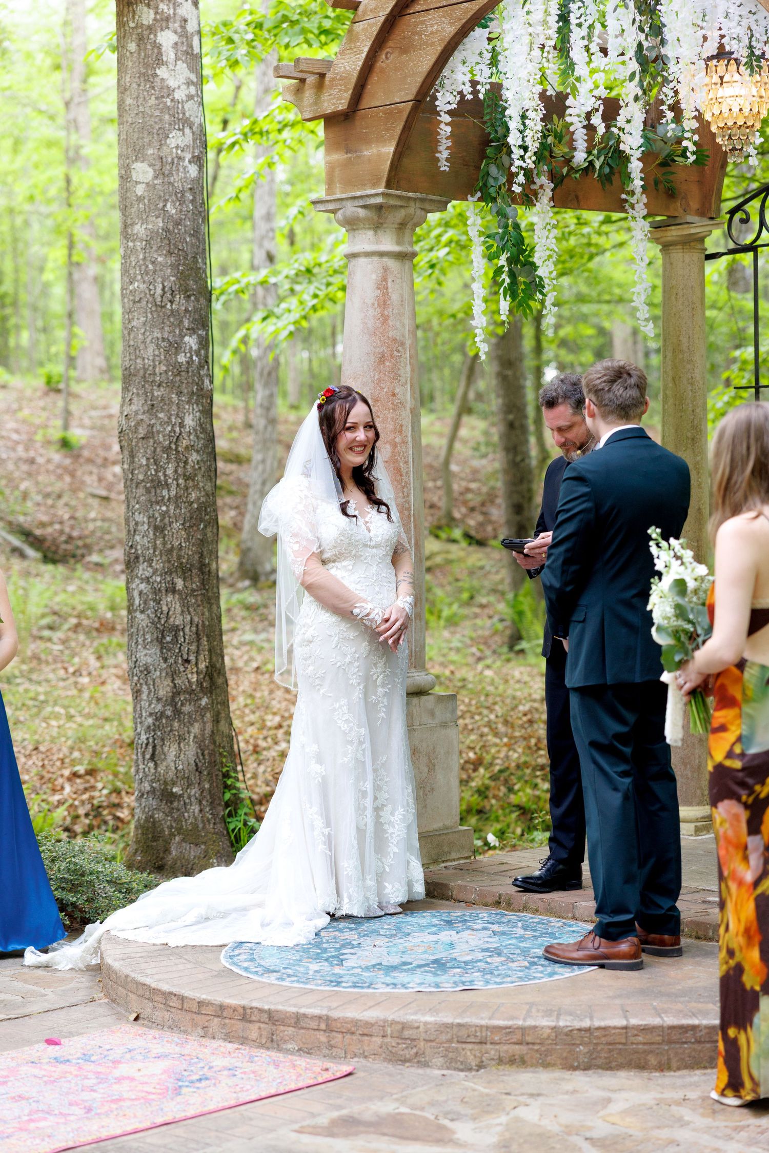 Bride smiling at groom during outdoor wedding ceremony under floral arch with wisteria.