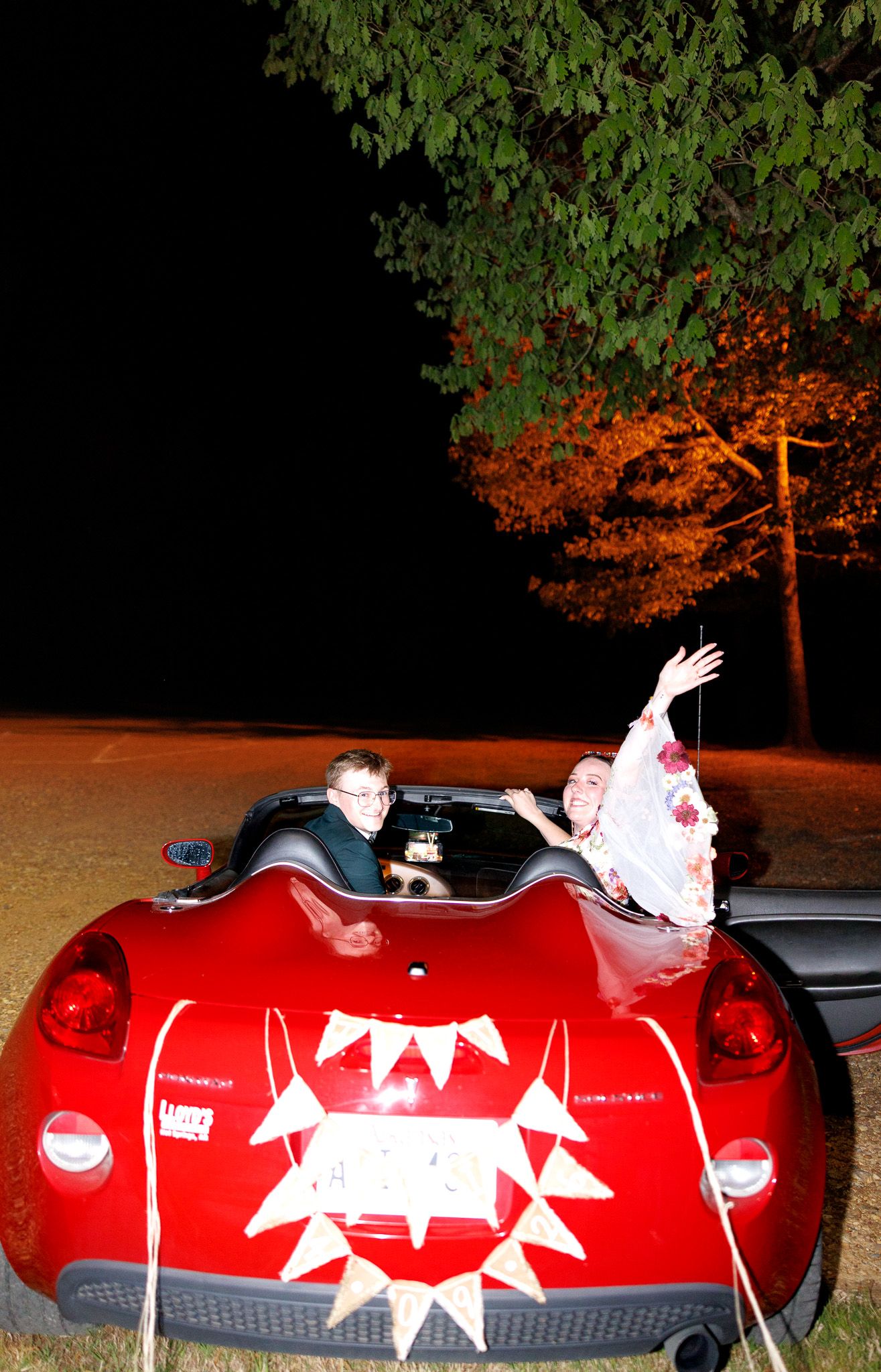 Two people wave from a red convertible sports car at night with illuminated trees in the background.