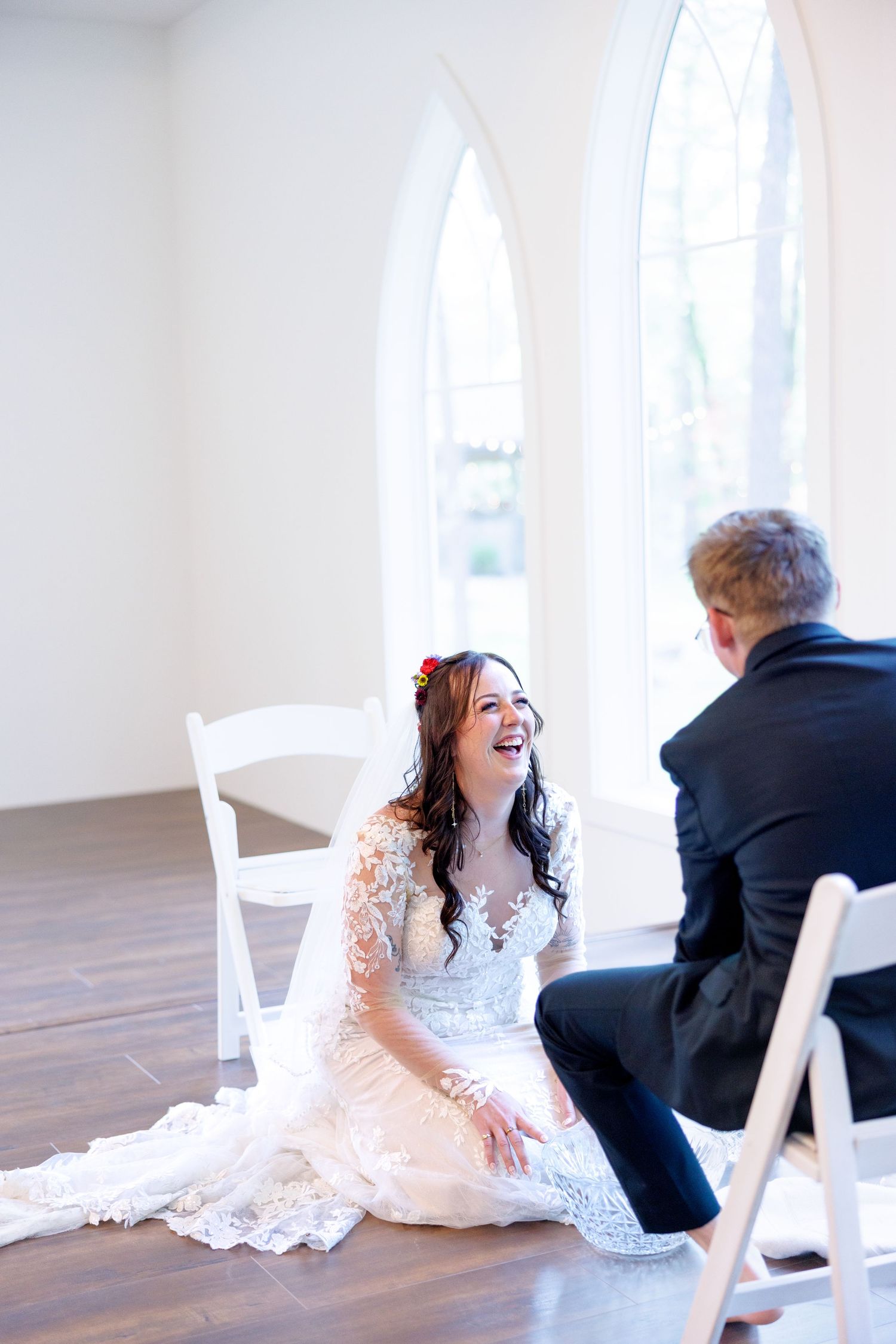 Bride in lace wedding dress laughing joyfully while sitting on floor with groom during intimate wedding moment.