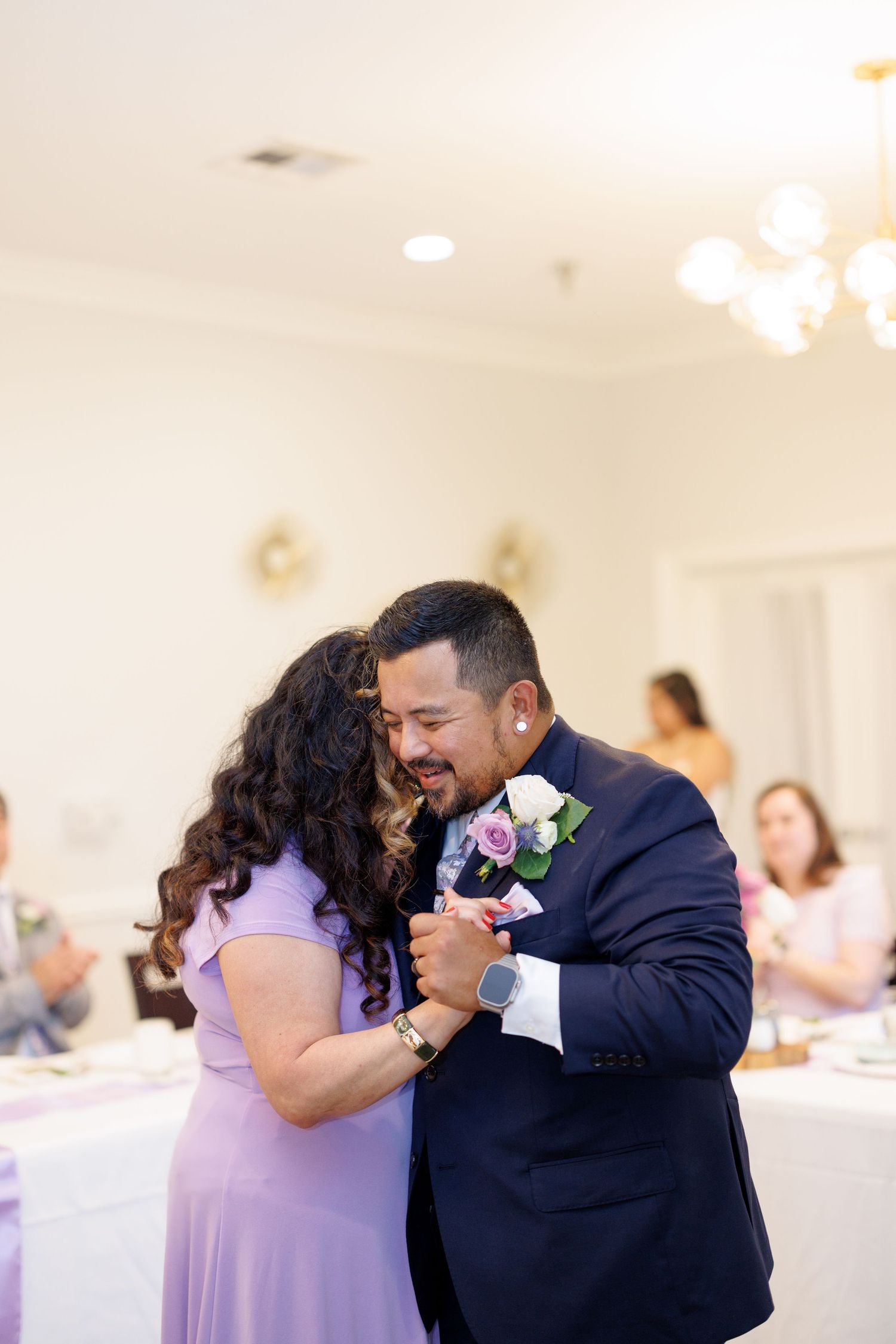 Groom in navy suit shares mother-son dance with woman in lavender dress at elegant wedding reception.