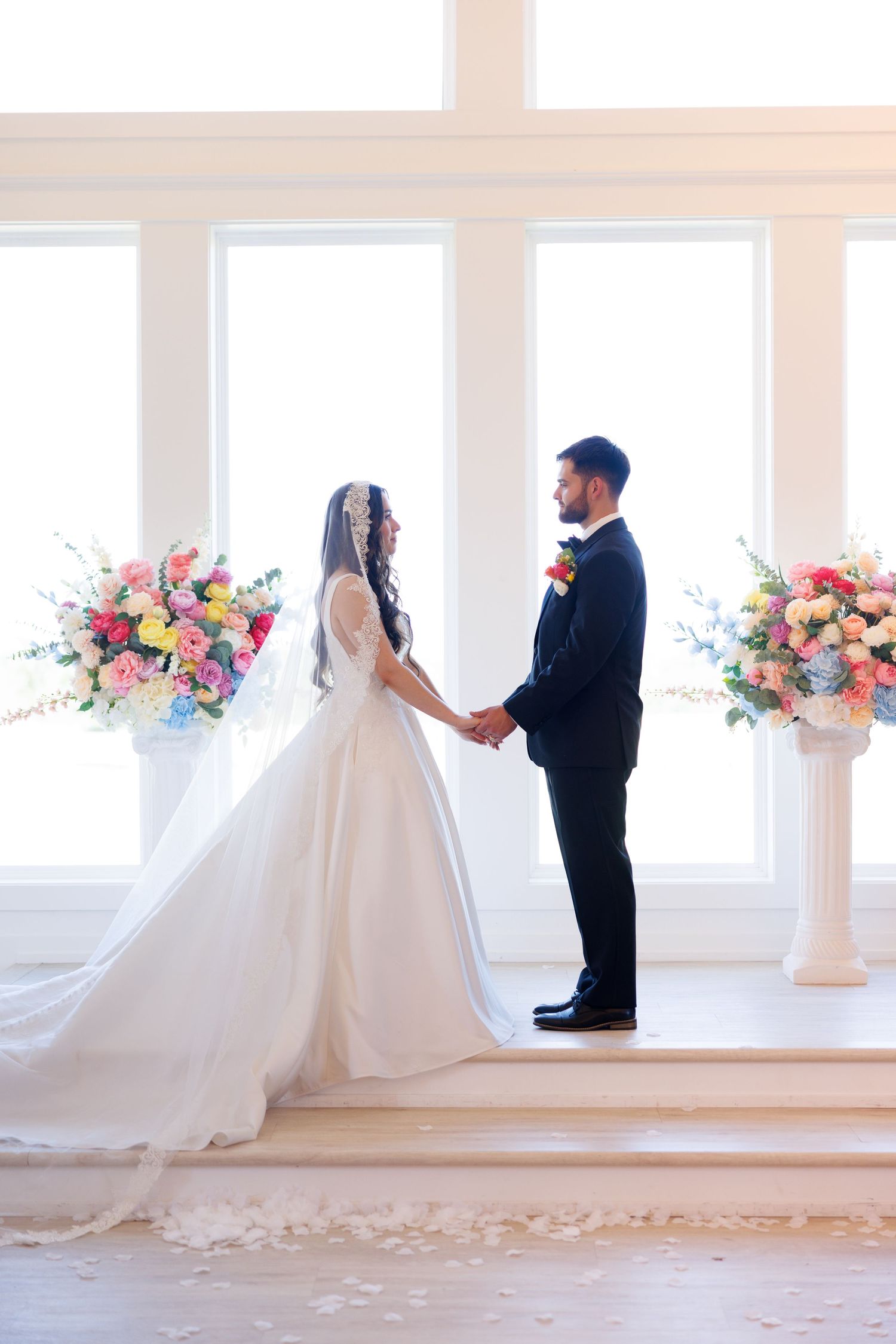 A wedding ceremony moment in front of large windows with colorful floral arrangements.