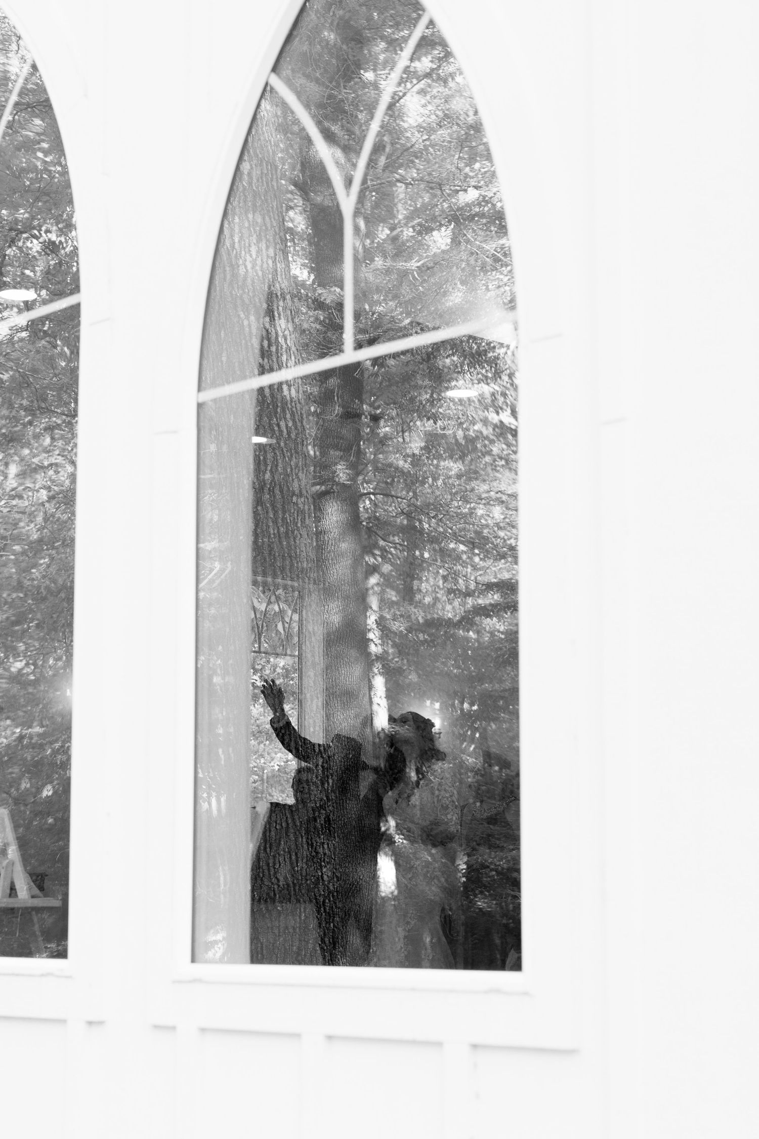 Black and white photo of two people visible through arched church windows, with trees reflected in the glass.