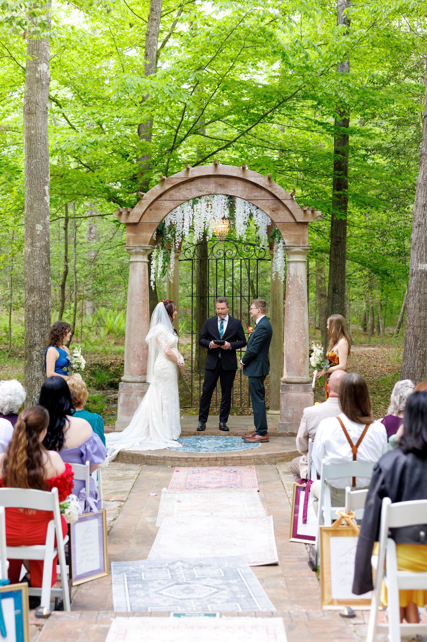 Bride and groom exchange vows under an ornate iron arch in a lush forest wedding ceremony.
