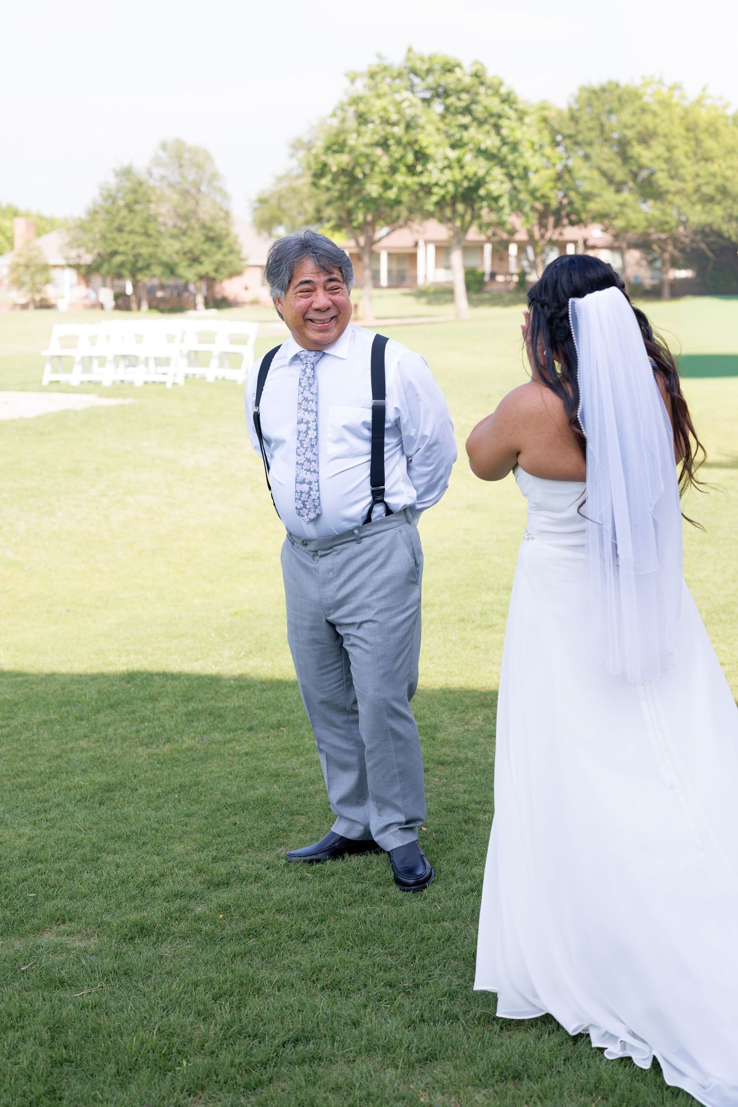 Smiling father in suspenders reacts to bride in white wedding dress and veil during outdoor first look ceremony.