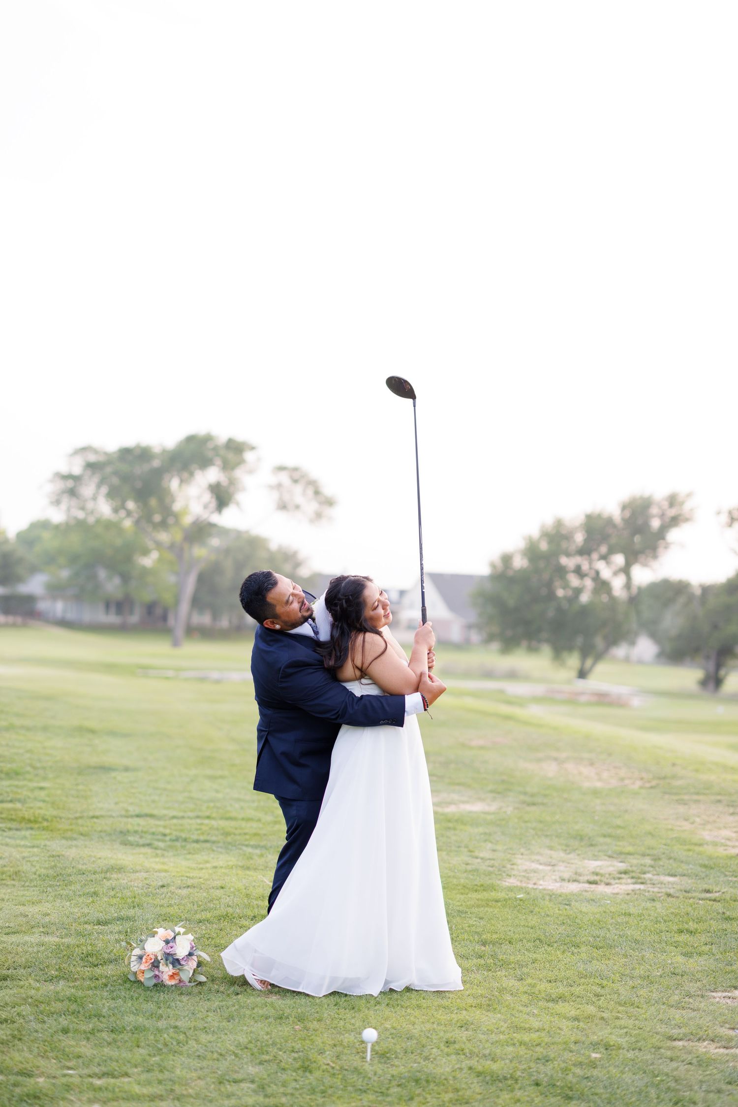 Bride and groom posing together swinging a golf club on a golf course during their wedding photo session.