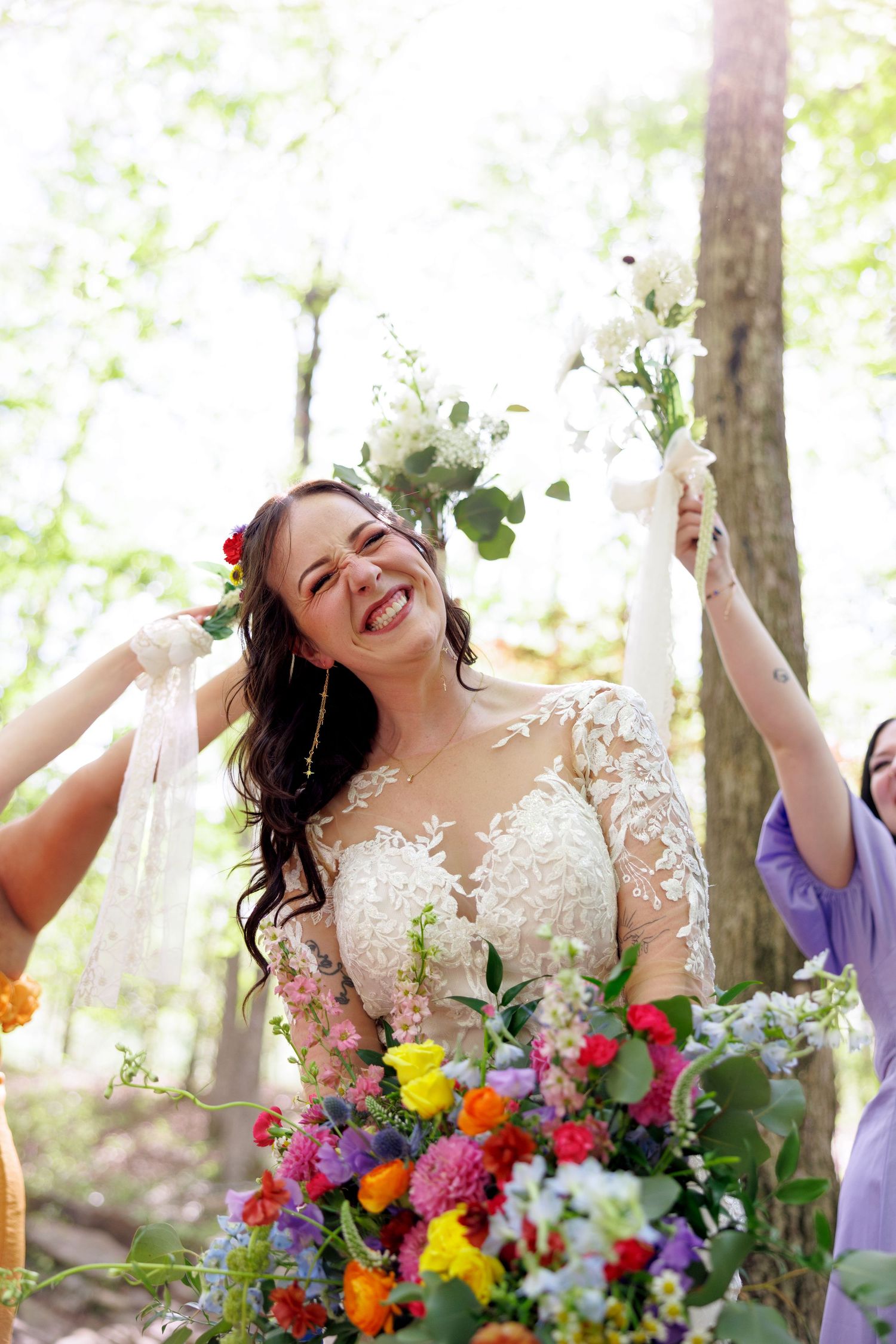 Joyful bride in lace gown laughs while holding colorful bouquet surrounded by bridesmaids in a bright forest setting.