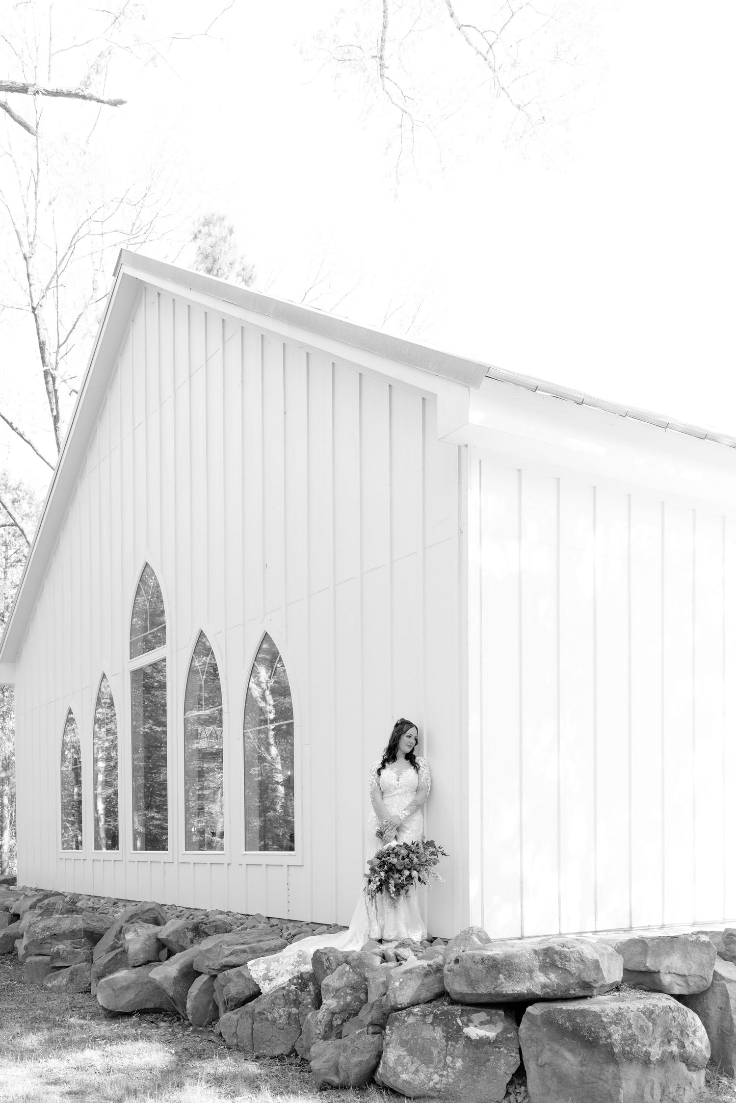 Black and white photo of a bride in a white gown holding a bouquet beside a white chapel with arched windows.